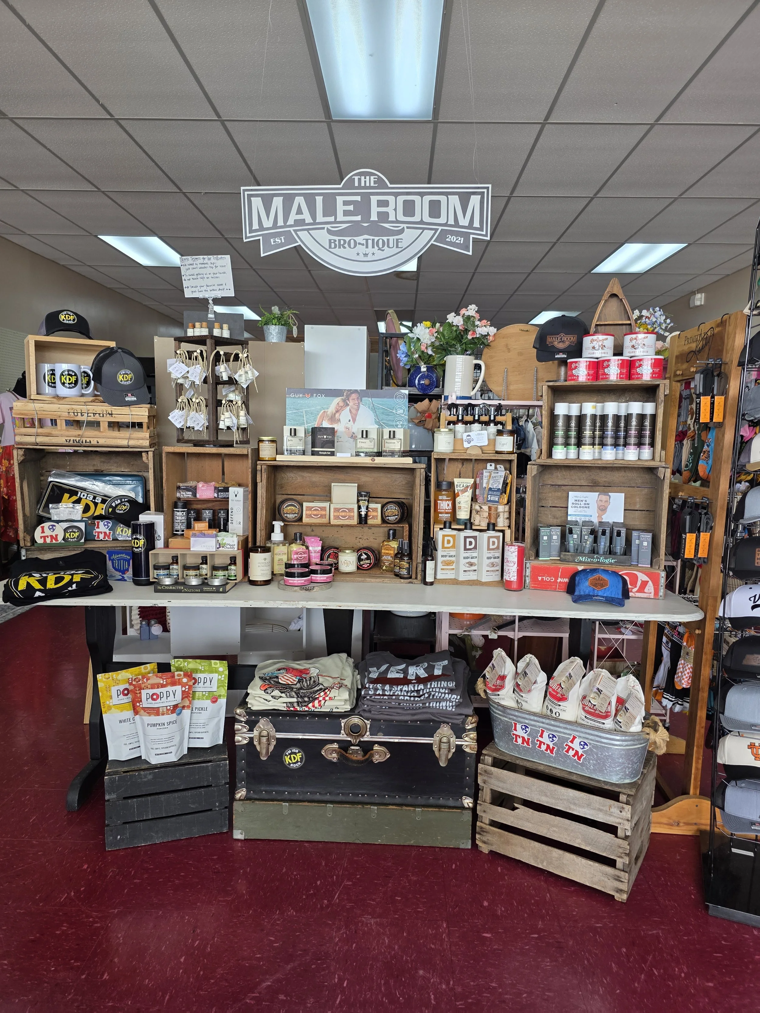 The Male Room Brotique retail display booth inside The Purple Iris Marketplace in Sparta, Tennessee, featuring men's graphic apparel, hats, grooming products, and artisan snacks.