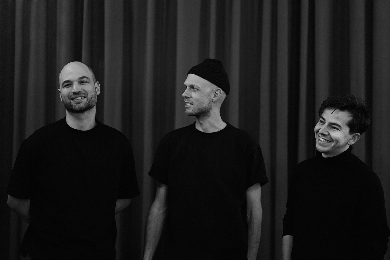 The three founders of coapp standing in front of a dark curtain, smiling and looking at each other in a black-and-white photo.