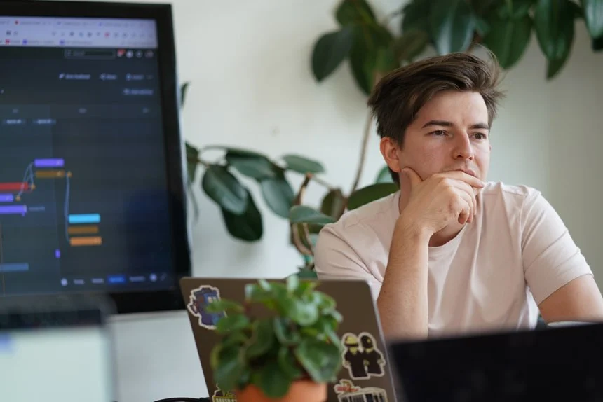 A young man working on a computer at a desk with a large monitor and laptop, surrounded by green plants.