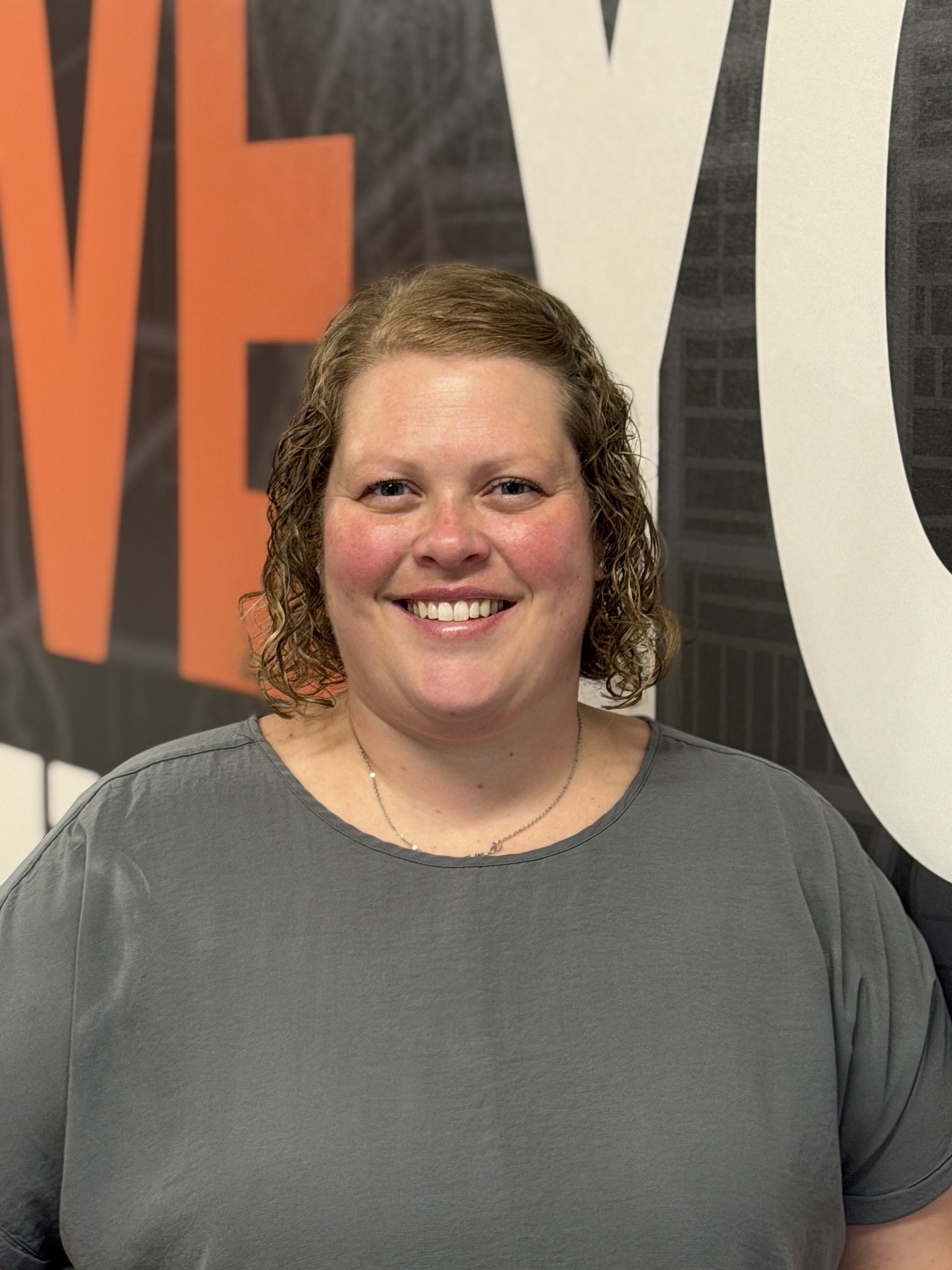 A woman with light skin and curly hair smiling in front of a black wall with large orange and white letters.