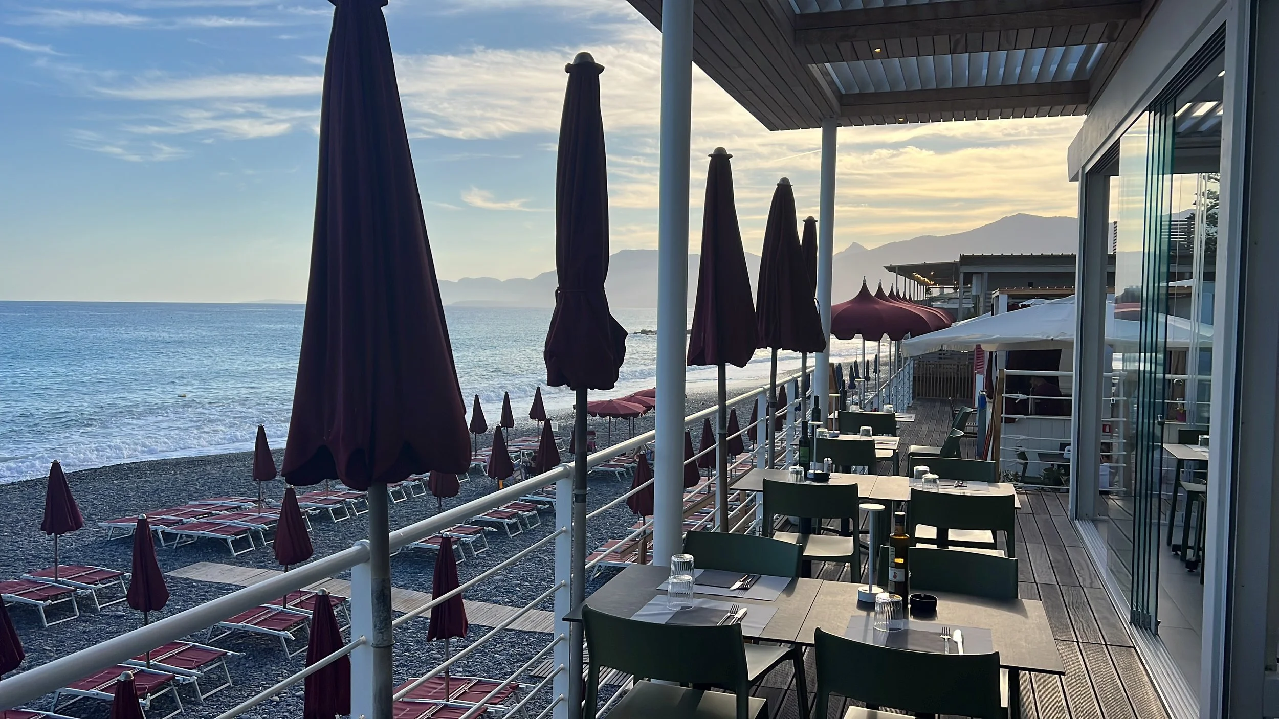 Balcone di un ristorante con tavoli e sedie, vista sulla spiaggia e sul mare di Bordighera, con ombrelloni chiusi e montagne sullo sfondo durante il tramonto.