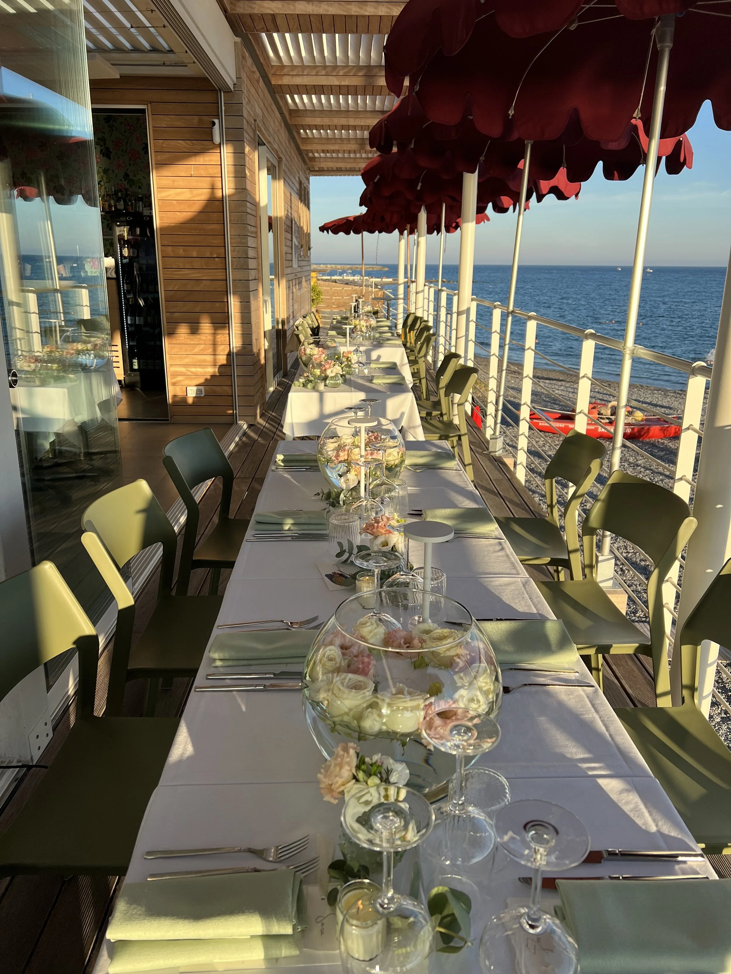 Tavola apparecchiata con fiori e bicchieri di vino, all'aperto sul mare con ombrelloni rossi e vista sull'oceano.