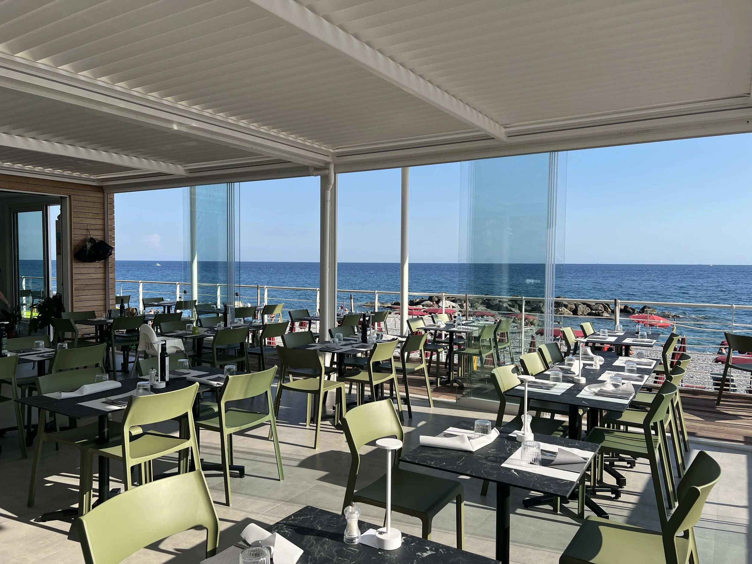 Terrazza di un ristorante con vista sul Mare di Bordighera, tavoli apparecchiati, sedie verdi e grandi vetri che mostrano il mare e il cielo azzurro.