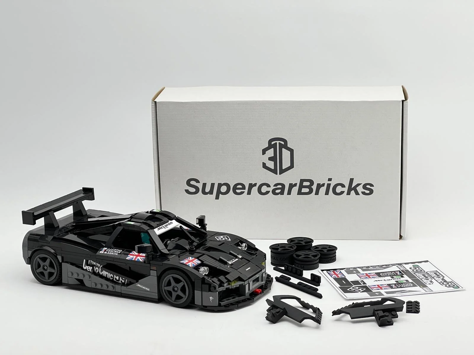 A LEGO model of a black race car with decals, a large rear wing, and decals of British flags, placed in front of a box labeled 3DSuperCarBricks, with LEGO pieces, stickers, and instructions nearby.