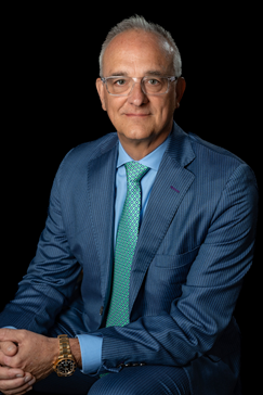 Rex Geveden wearing a blue suit, blue shirt and green tie in front of a black background.