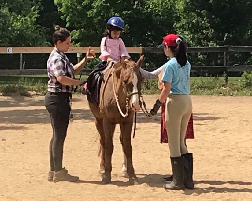 A young girl wearing a pink shirt and a helmet riding a horse, with two women on either side guiding and supporting her in an outdoor riding arena.
