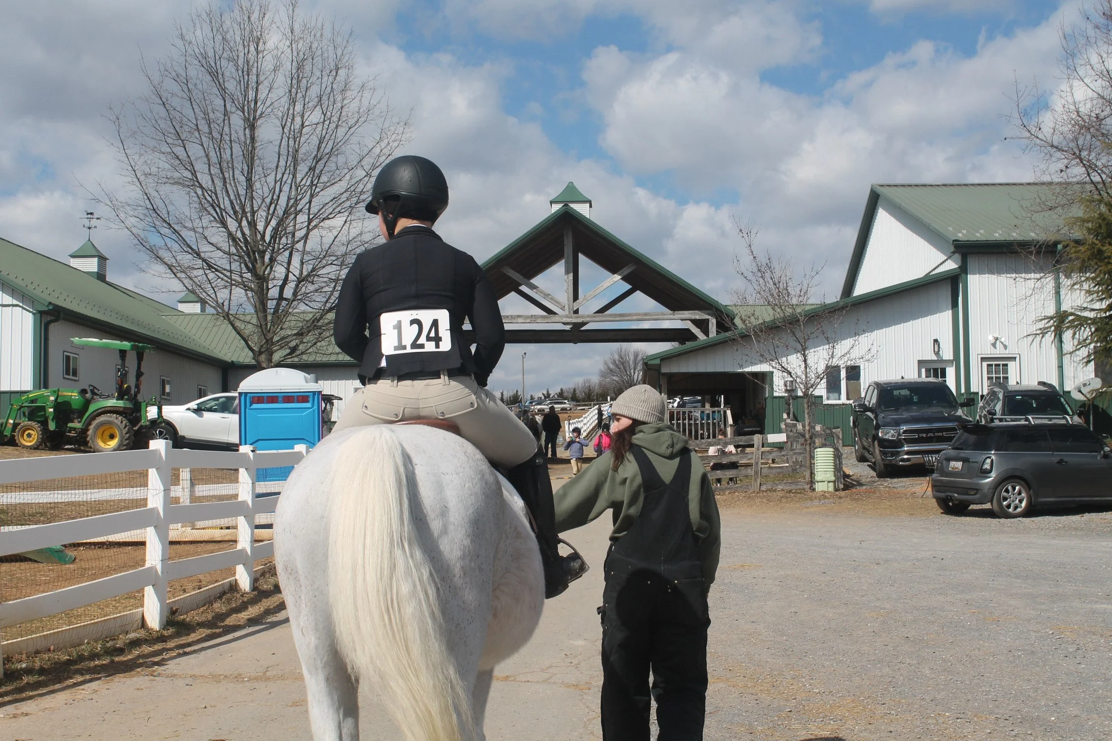 A person wearing a black helmet and matching jacket is riding a white horse, with a number 124 pinned to their back. Another person, dressed in a green hoodie and black pants, is walking beside the horse, holding its reins. The scene takes place outd