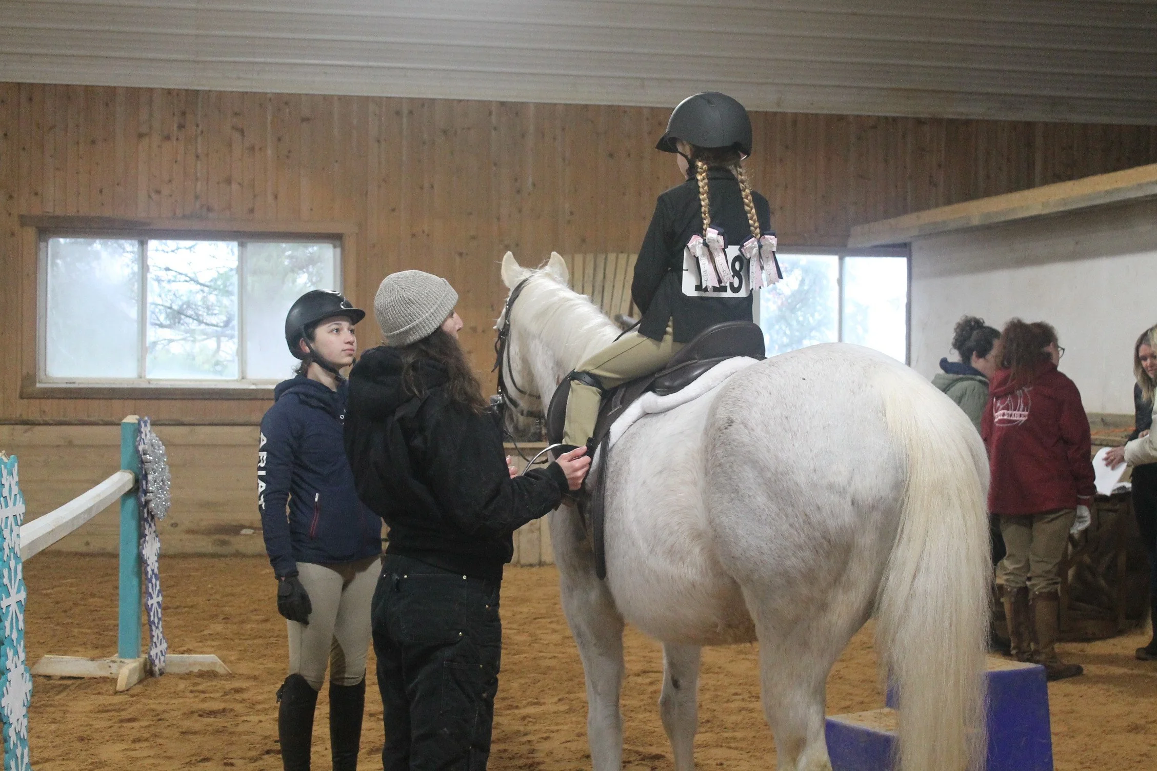 A girl in riding gear sitting on a white horse inside an indoor riding arena, with two women and other people nearby.