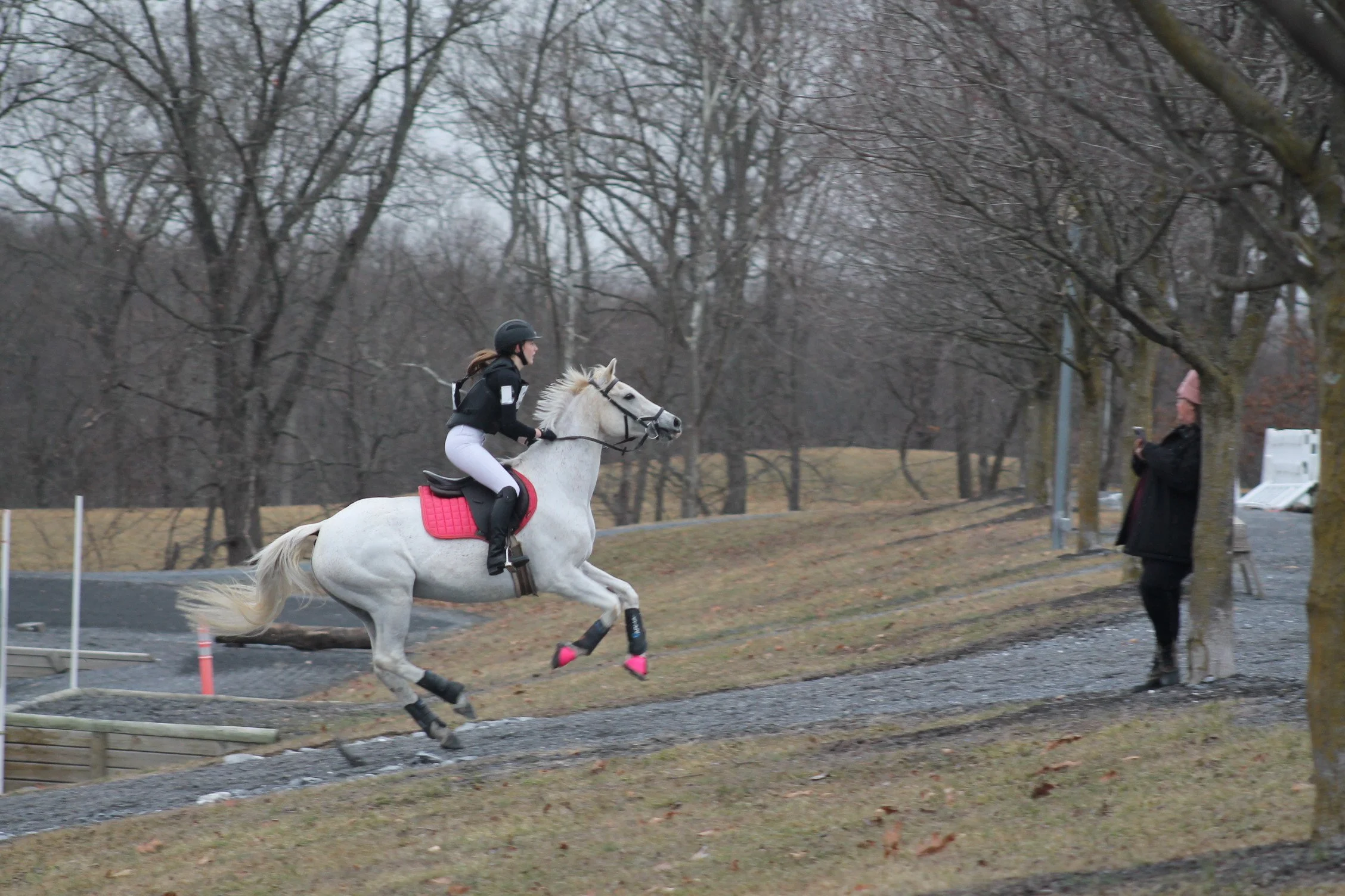 A female equestrian riding a white horse during a jump over a low obstacle in an outdoor setting on a cloudy day, with a person taking photos nearby.