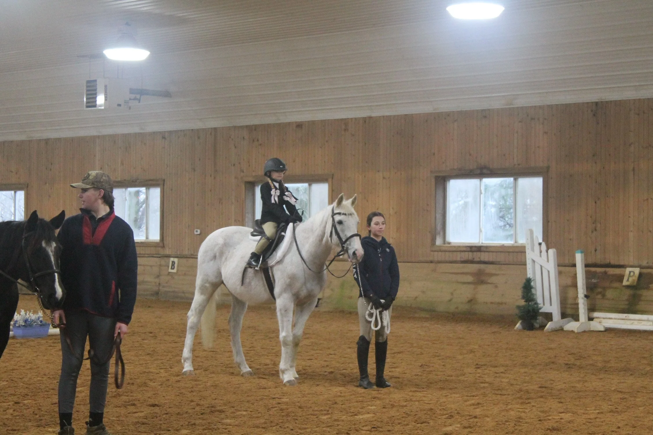 A young girl riding a white horse in an indoor riding arena, with two women standing nearby and a small jump obstacle in the background.