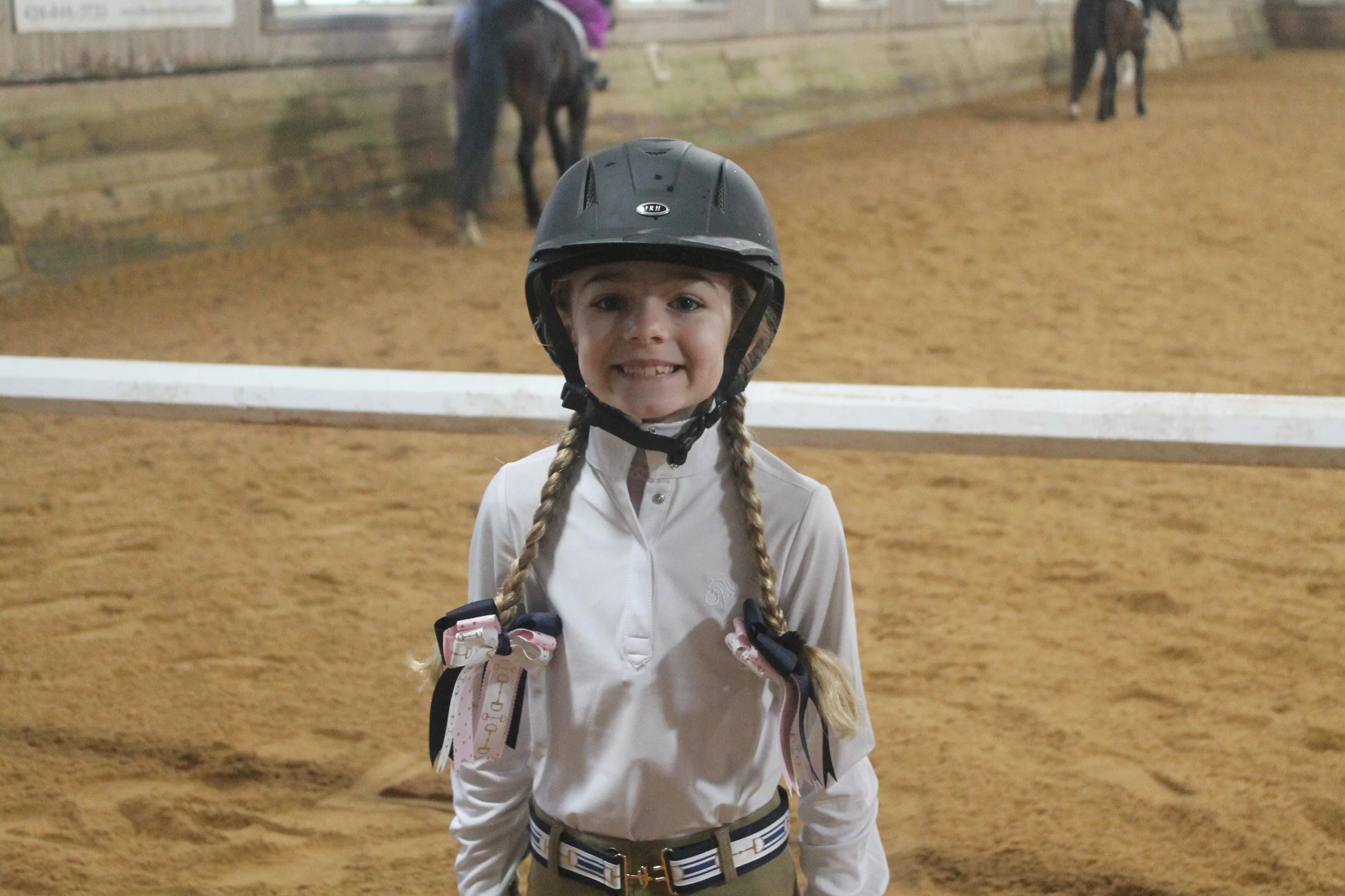 Young girl smiling at the camera on a horse riding arena, wearing a helmet, white shirt, and riding gear, with horses in the background.