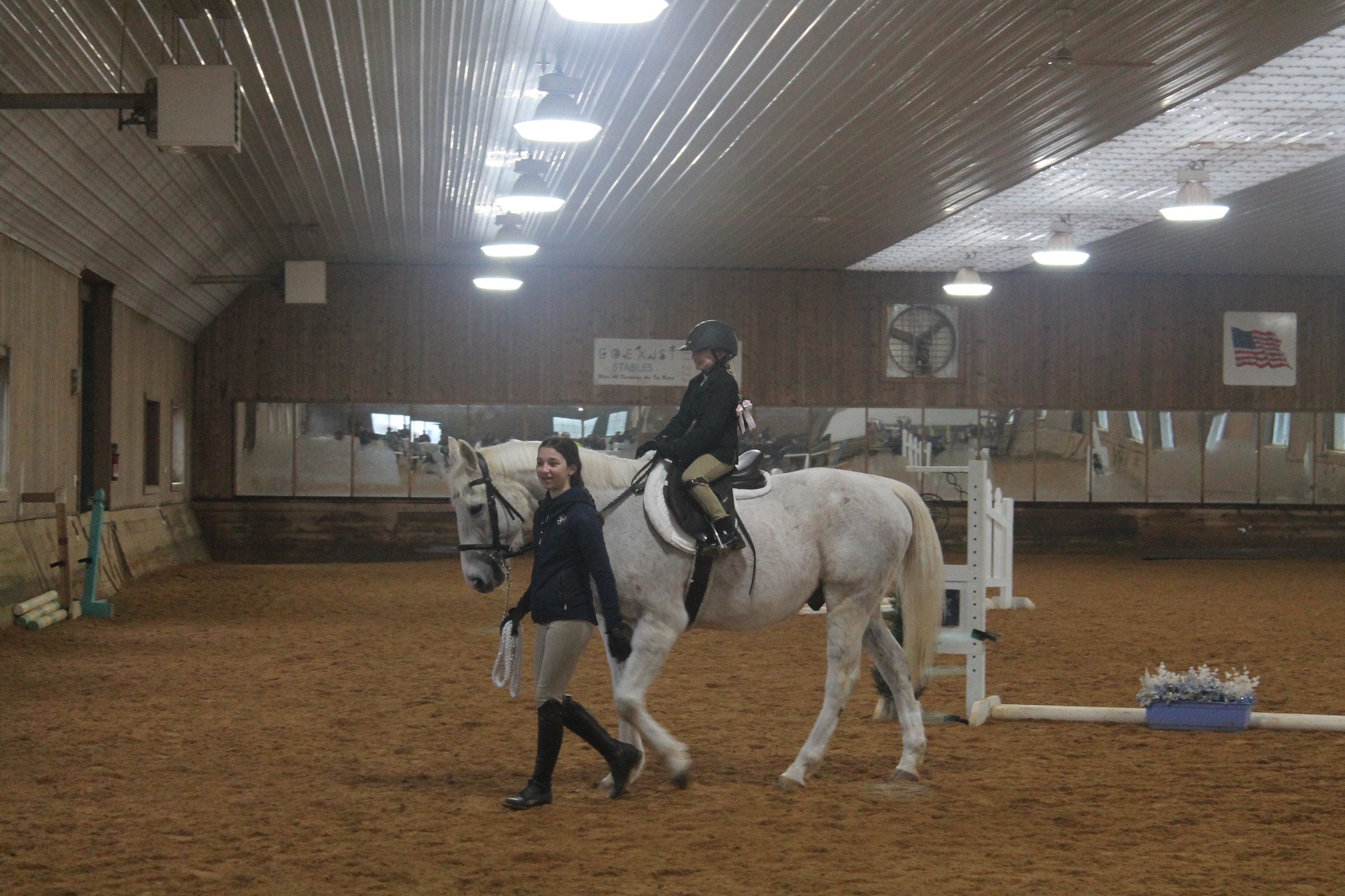 In an indoor horse riding arena, a young girl in riding gear is seated on a white horse, with a woman walking alongside holding the horse's bridle. The arena has a sand floor, wooden walls, ceiling lights, and American flags.