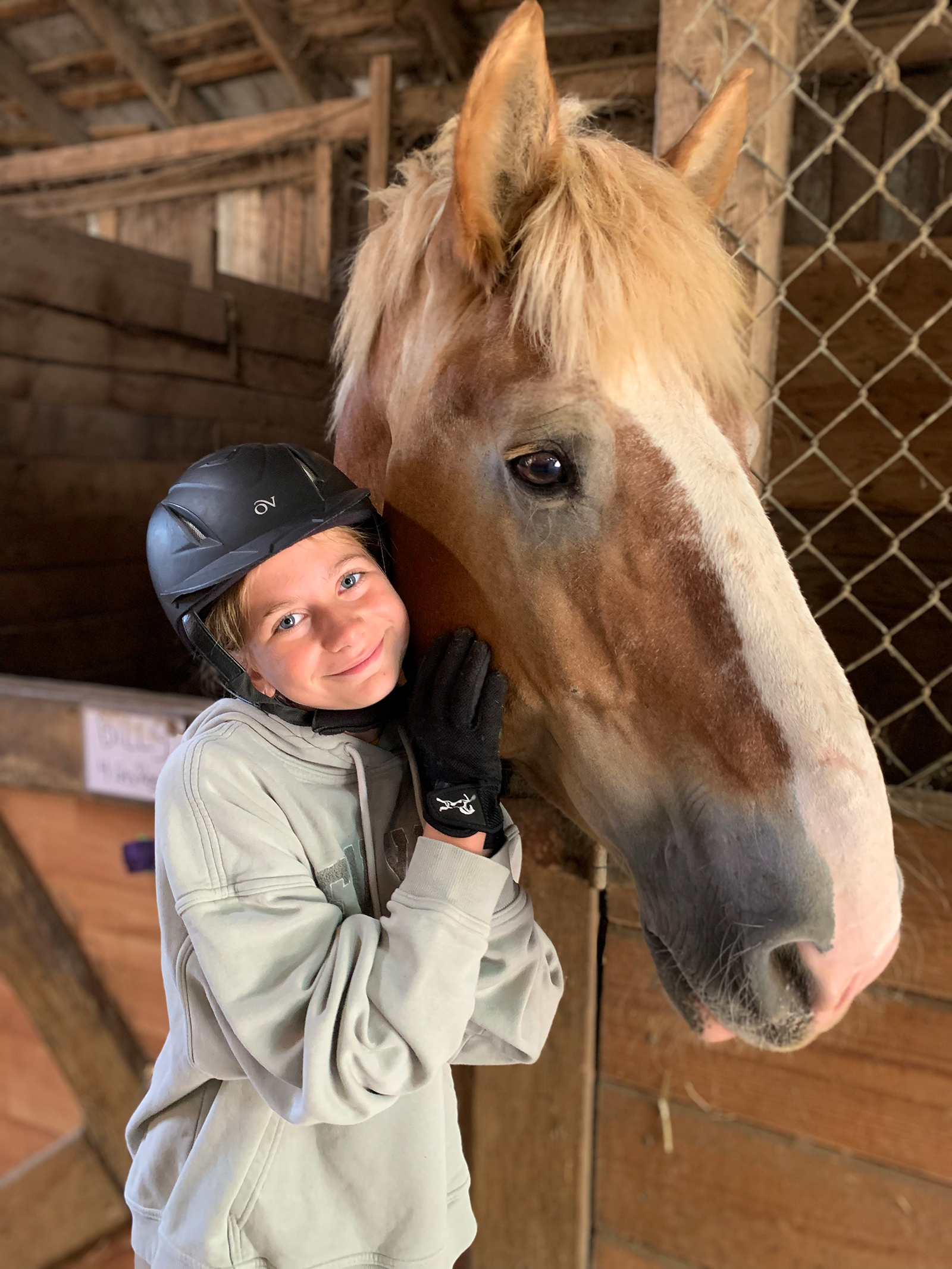 A young girl with a black riding helmet, hoodie, and gloves cuddling a light brown horse with a white blaze on its face inside a wooden stable.