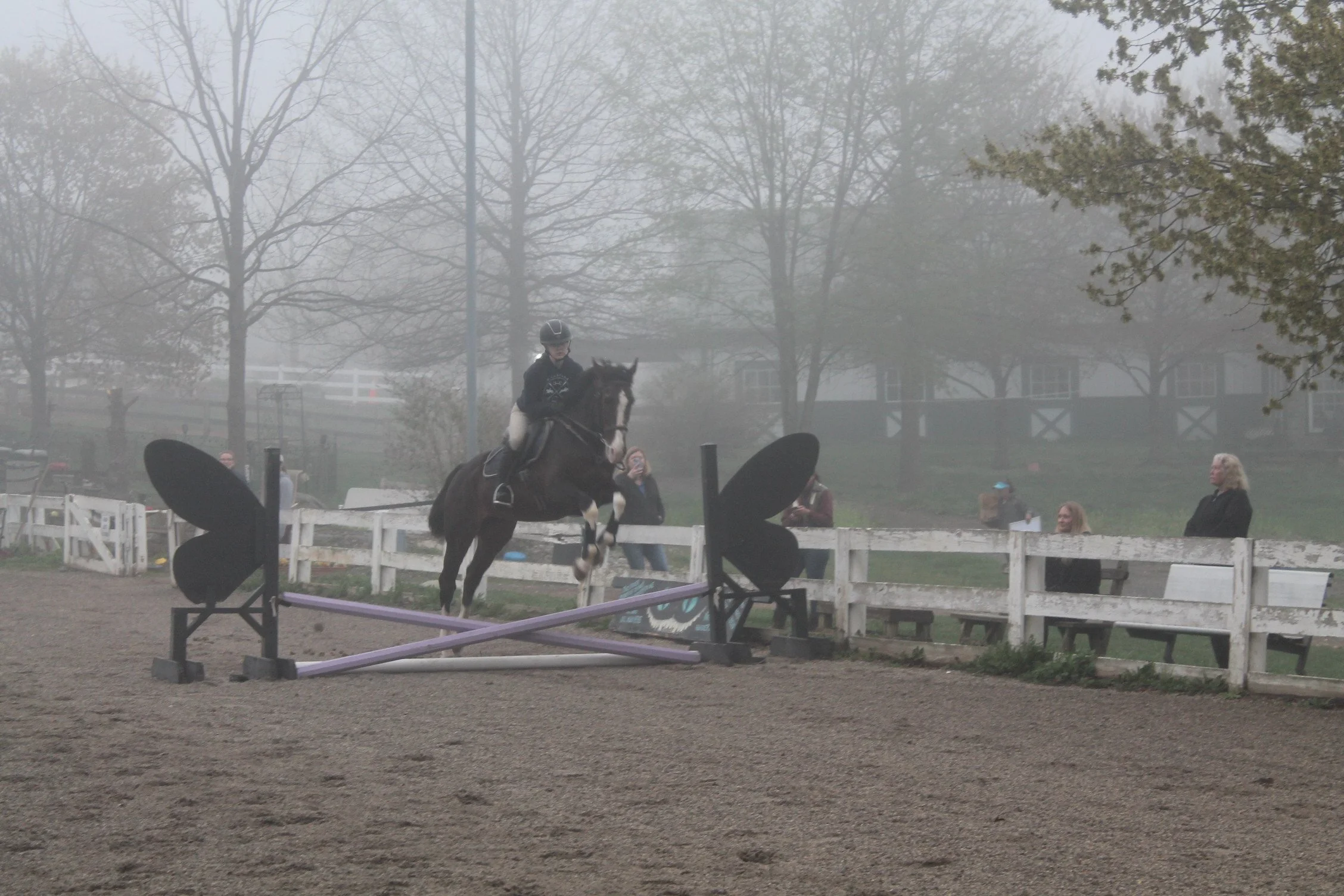 A person riding a horse jumps over an obstacle in an outdoor equestrian arena on a foggy day.
