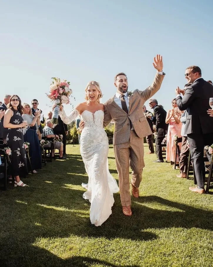 Bride and groom celebrating as they walk down the aisle at their outdoor wedding, surrounded by guests clapping and smiling.