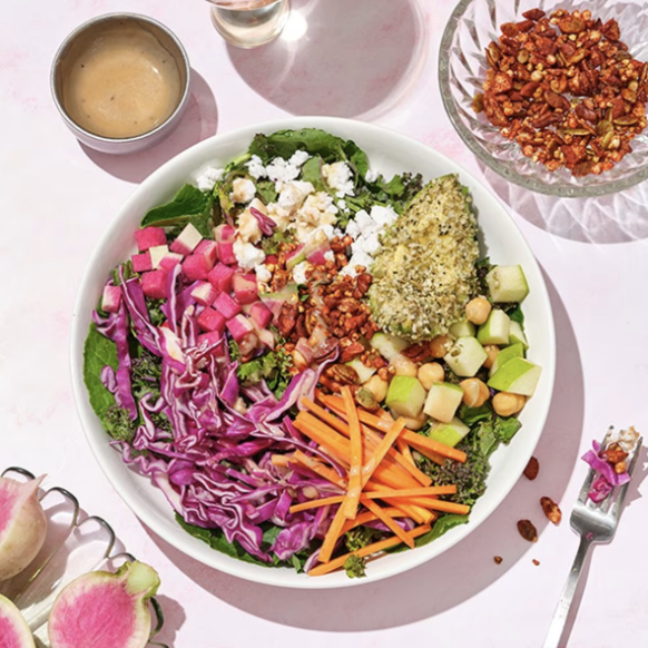 A woman with long dark hair smiling in a modern kitchen, leaning on a white marble countertop. She is wearing a white cardigan and jeans, with a glass of water and a bowl of lemons on the counter. There are flowers in the background.