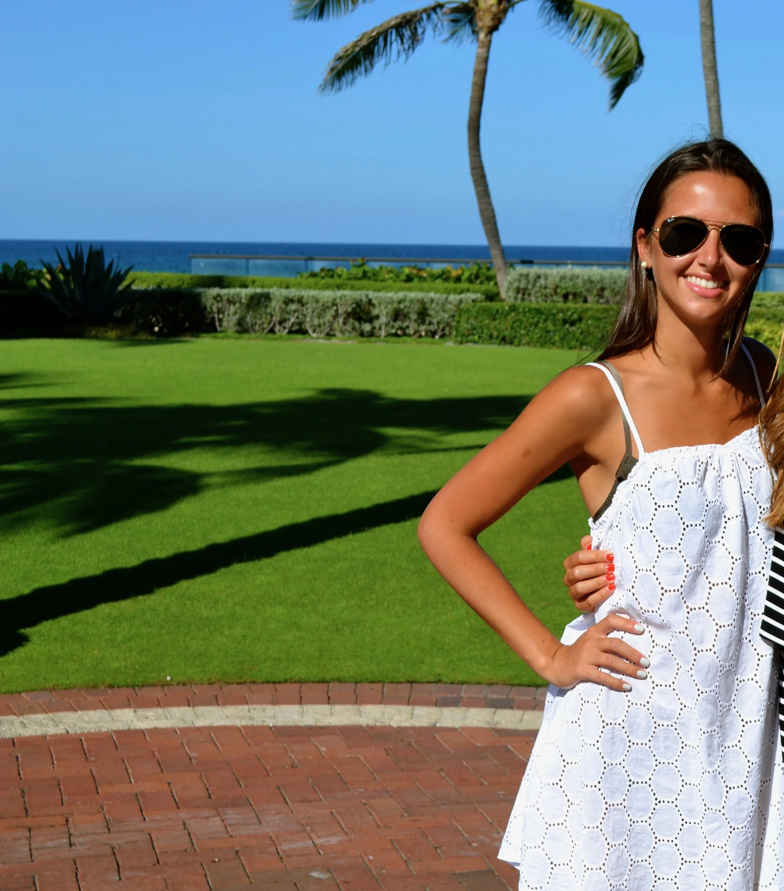 A woman in sunglasses and a white summer dress stands on a brick path with lush green grass, palm trees, and the ocean in the background.