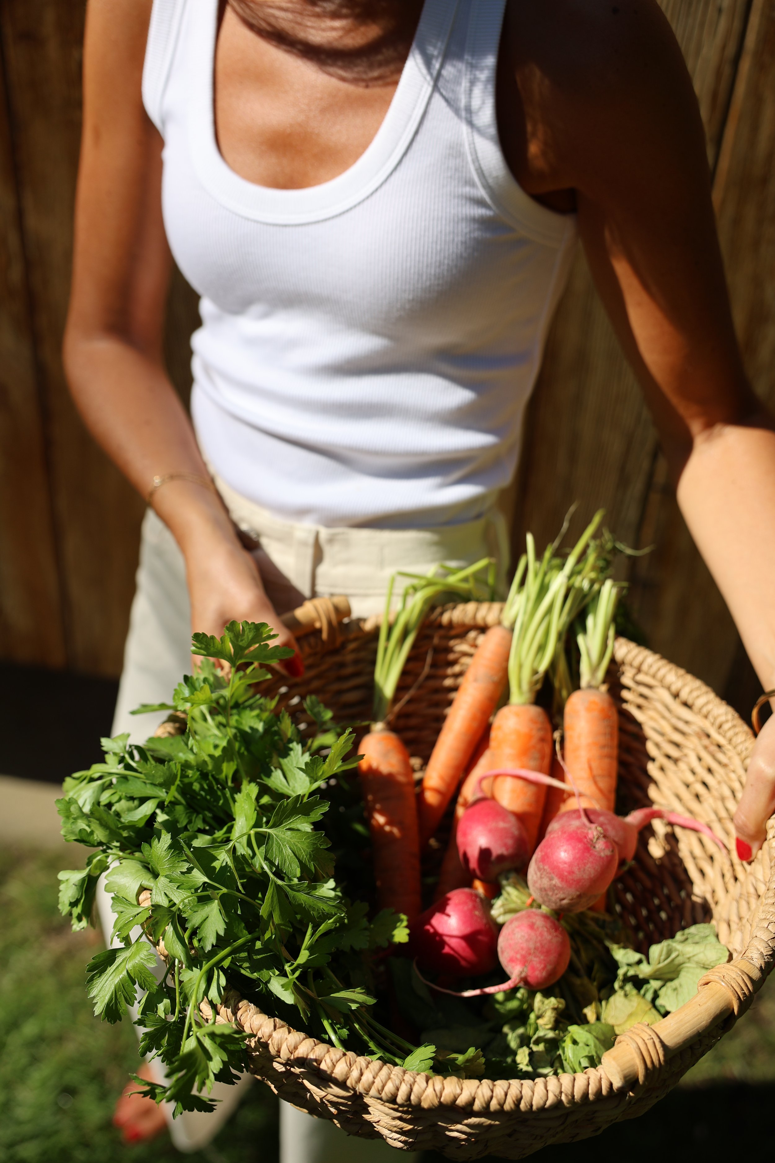 A woman holding a basket of freshly harvested vegetables, including carrots, radishes, and parsley, outside in the sunlight.