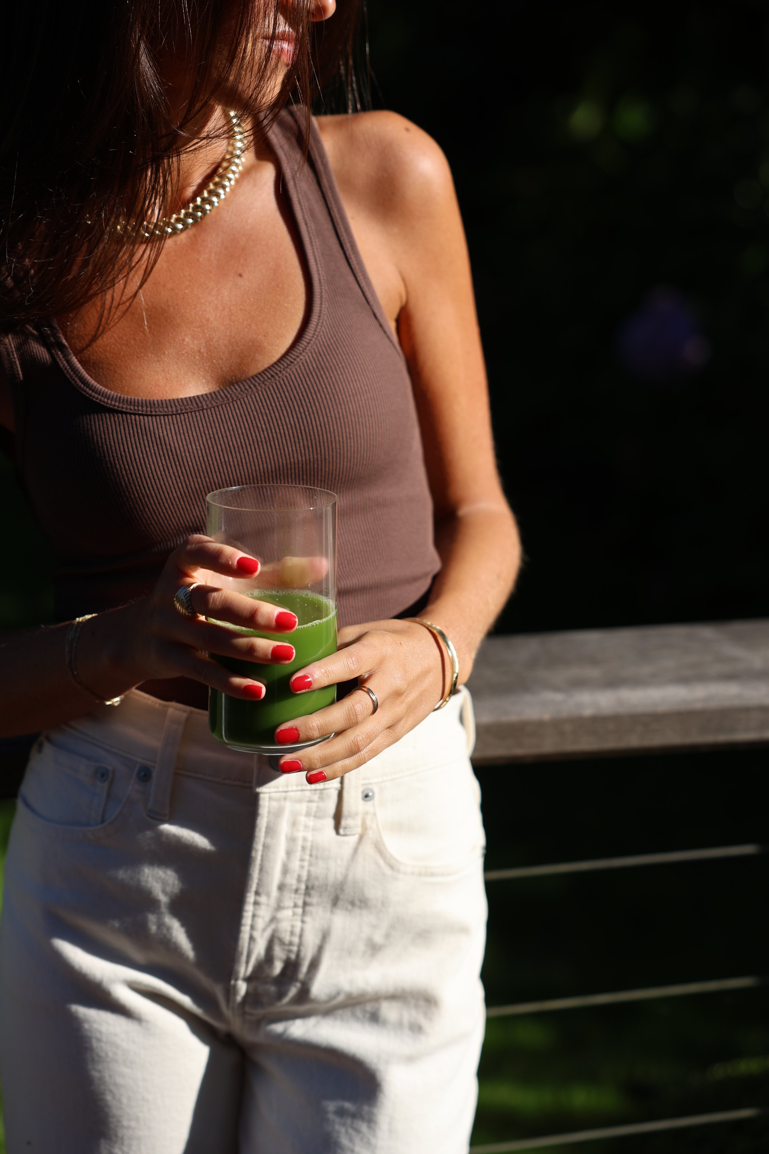 A woman in a brown sleeveless top and white pants holding a glass of green juice outdoors, with sunlight highlighting her shoulder, wearing jewelry including a pearl necklace and rings.