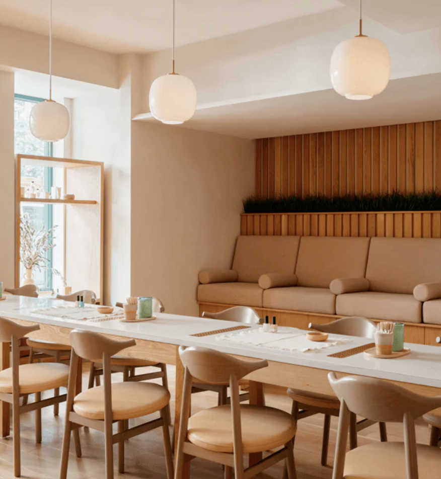A cozy dining area with a long white table, light wood chairs, and a beige sofa against a wooden-paneled wall with greenery. Three white pendant lights hang from the ceiling, and a window with natural light is present.