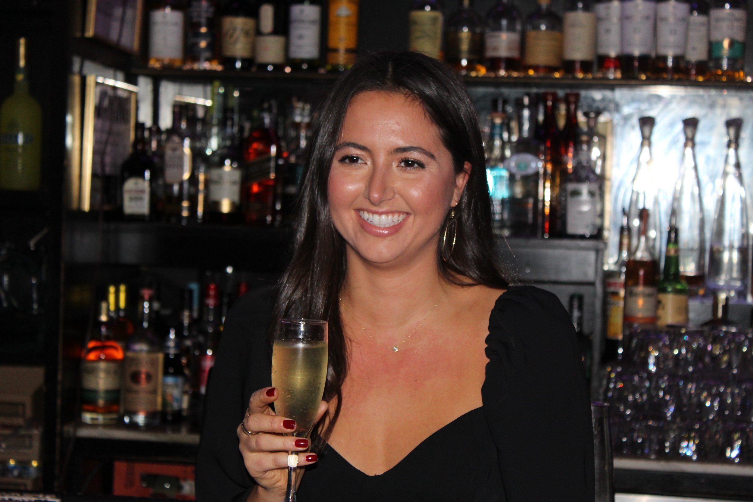 A woman with long dark hair, wearing a black top, holding a glass of champagne, smiling at a bar with various bottles of alcohol in the background.