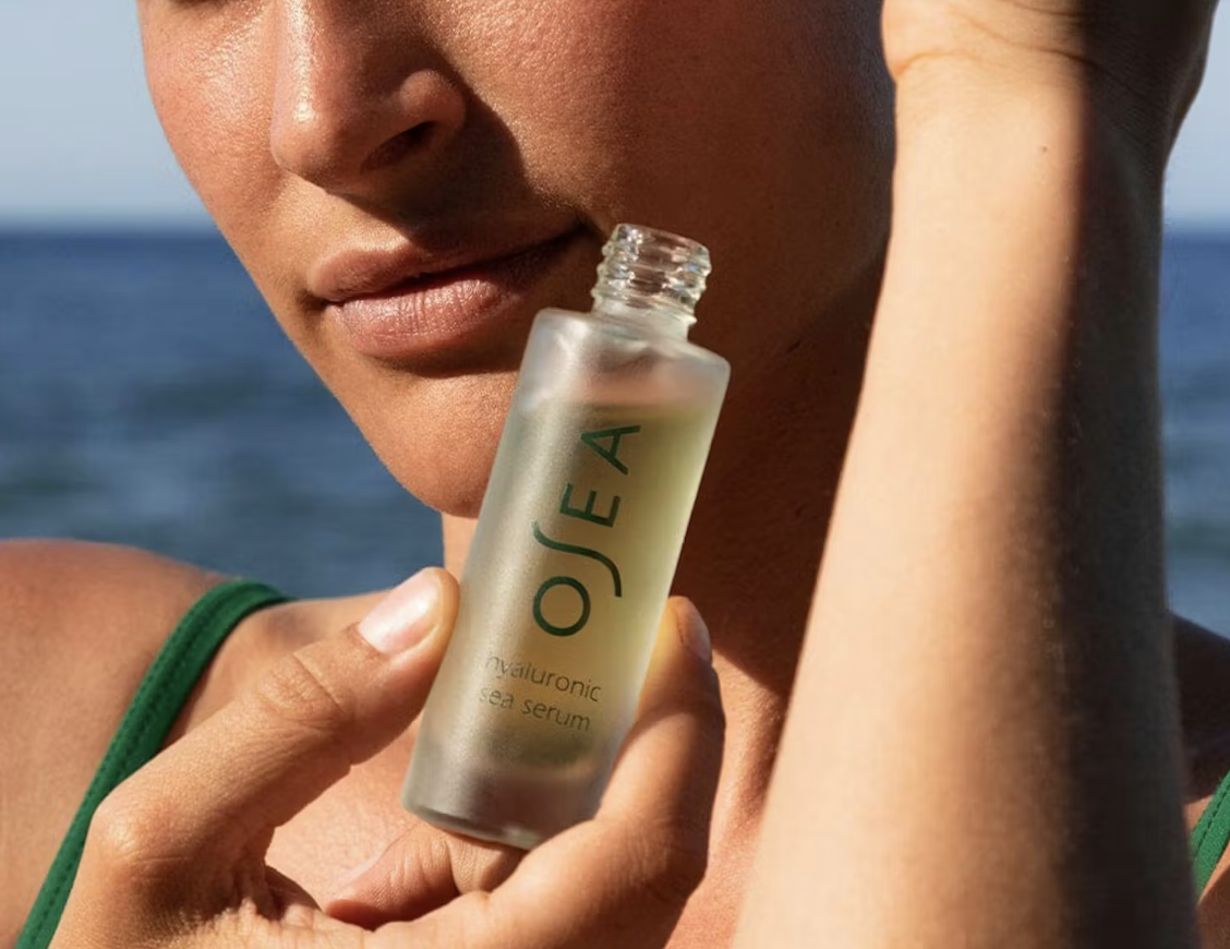 A woman with shoulder-length brown hair smiling and laughing outdoors on a sunny day, holding a glass vase with pink and green hydrangea flowers, standing on a wooden deck with a lush green background.