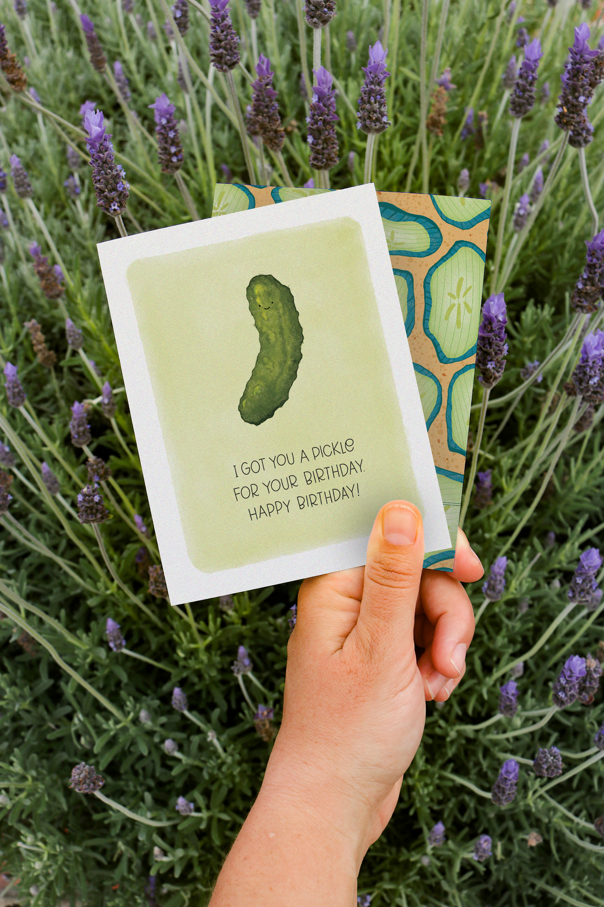 Hand holding "I Got You a Pickle for Your Birthday" greeting card by The Noble Paperie against a lavender field background