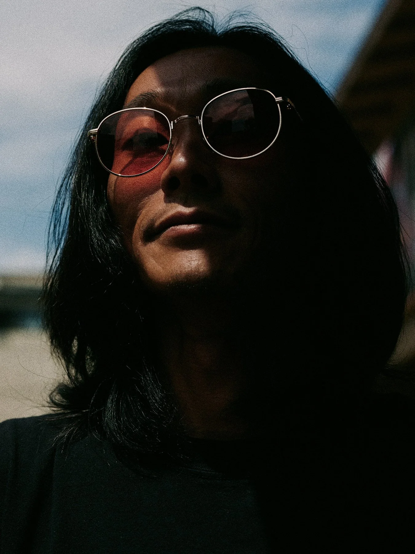 Close-up of a woman with black hair wearing round sunglasses, with sunlight casting shadows on her face, outdoors during daytime.