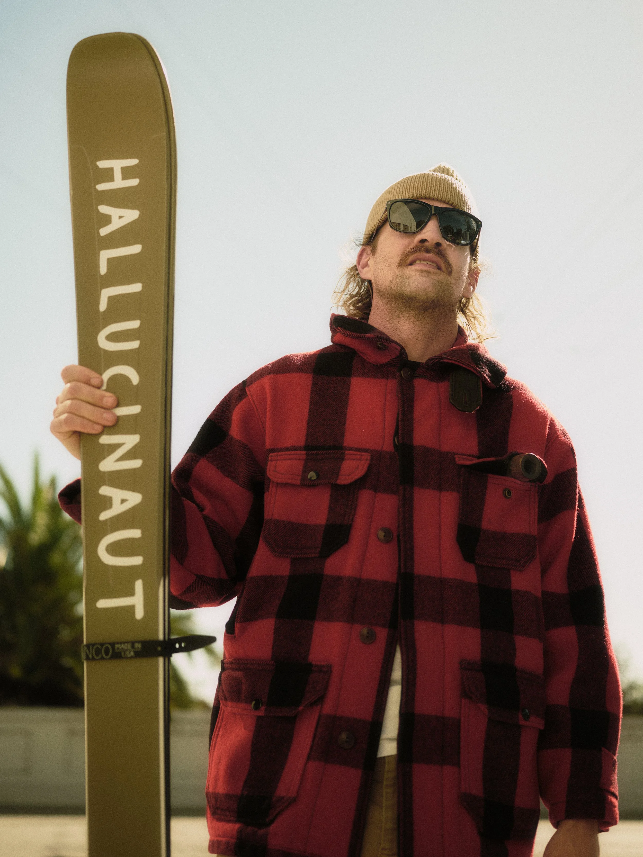 A man with sunglasses, a beige beanie, and a red plaid jacket holding a yellow snowboard with the words 'HALLUCINAUT' written on it.