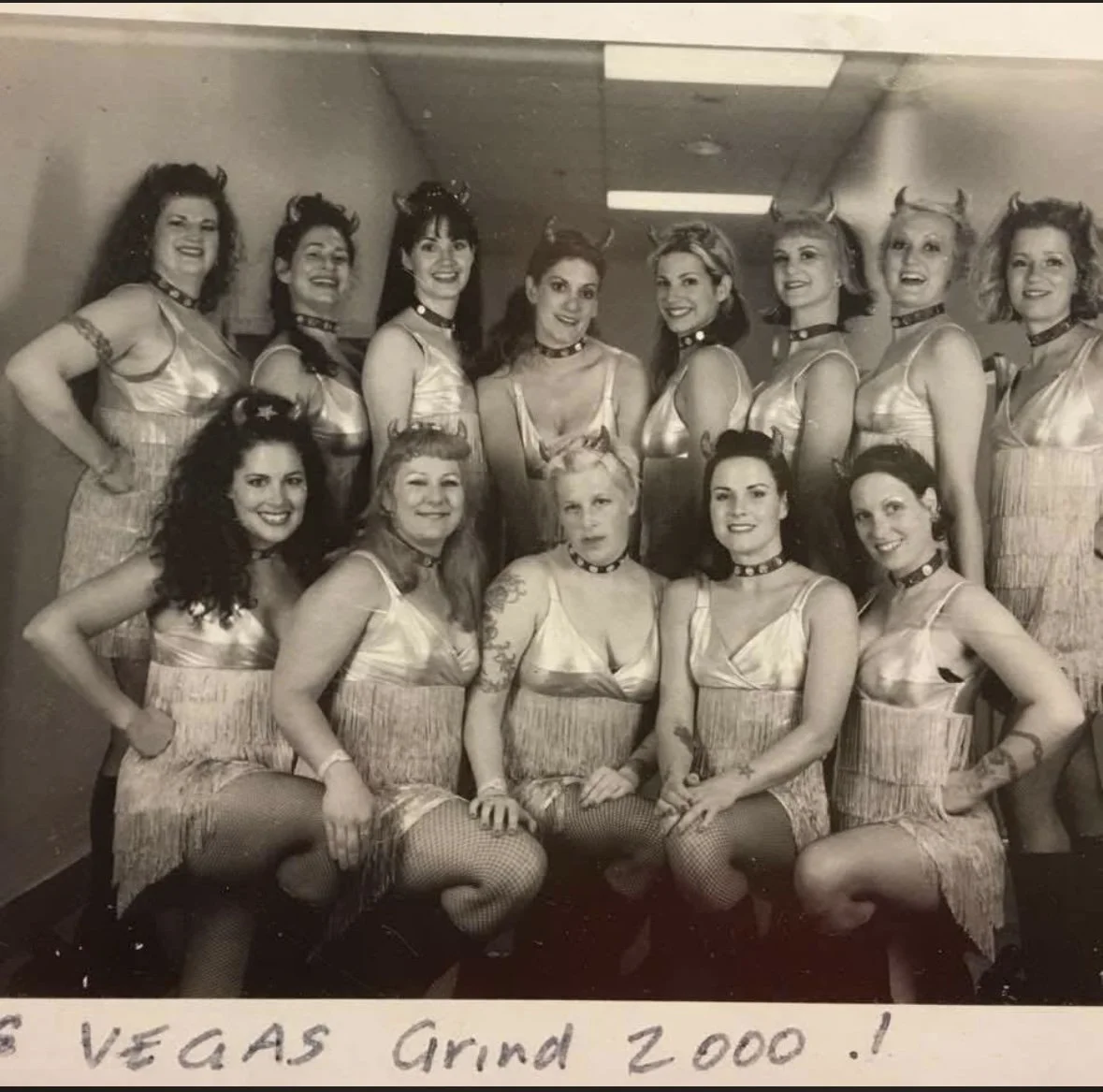Vintage black-and-white group photo of the Devil-Ettes performance troupe backstage at Vegas Grind 2000 in San Francisco, wearing matching fringe dresses, devil horns, fishnet stockings, and smiling before a burlesque-inspired performance.