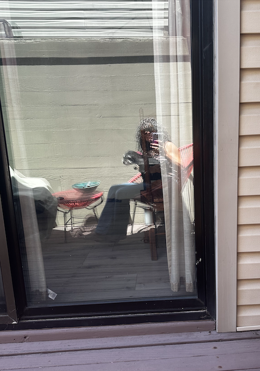 Reflection of a woman with curly hair sitting on a sunny San Francisco patio, photographed through a window, creating a quiet, nostalgic self-portrait about home, reflection, and nearly three decades of living in San Francisco.