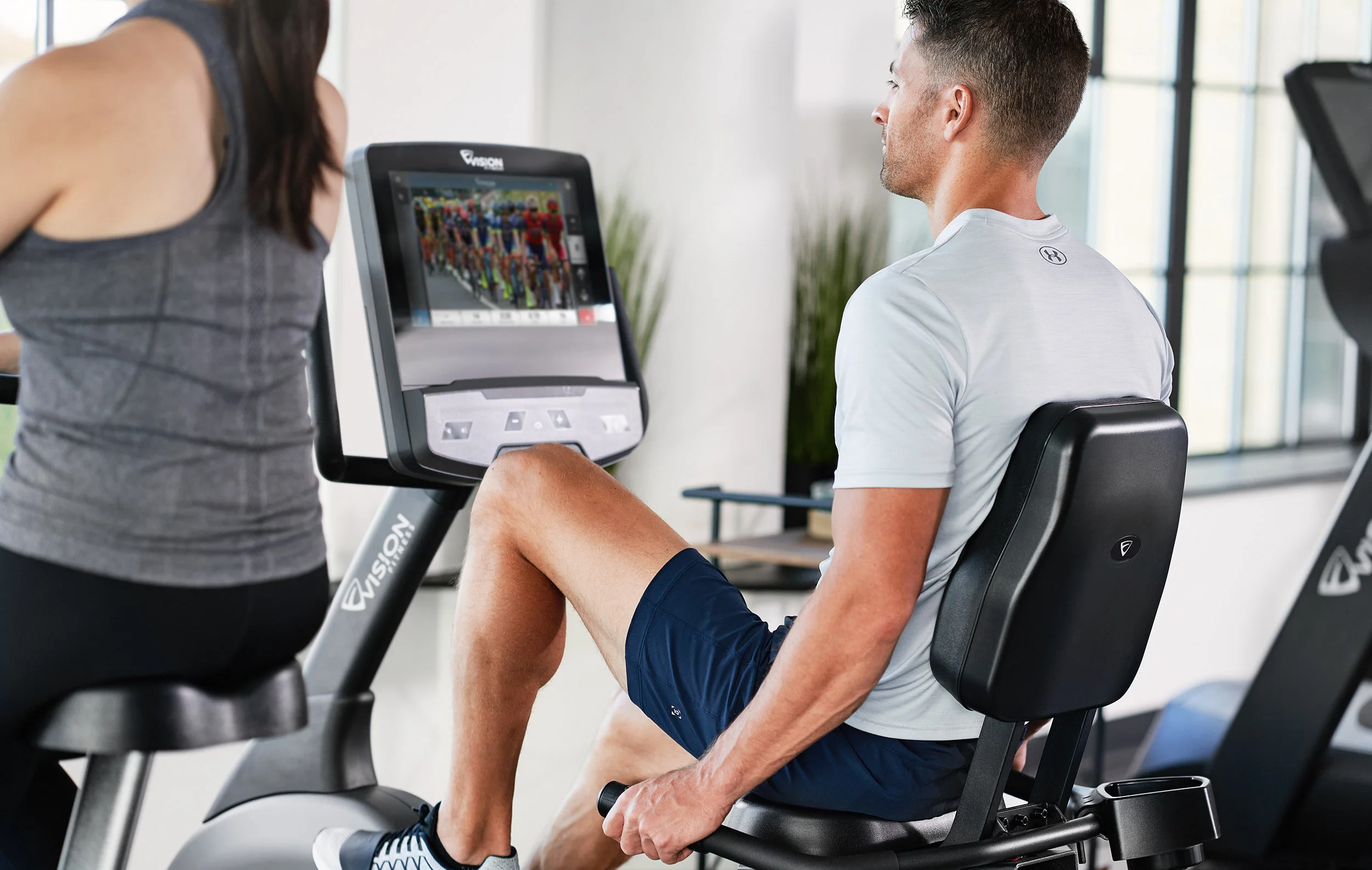 A man on a stationary exercise bike watching a screen in a gym, with a woman standing next to him.