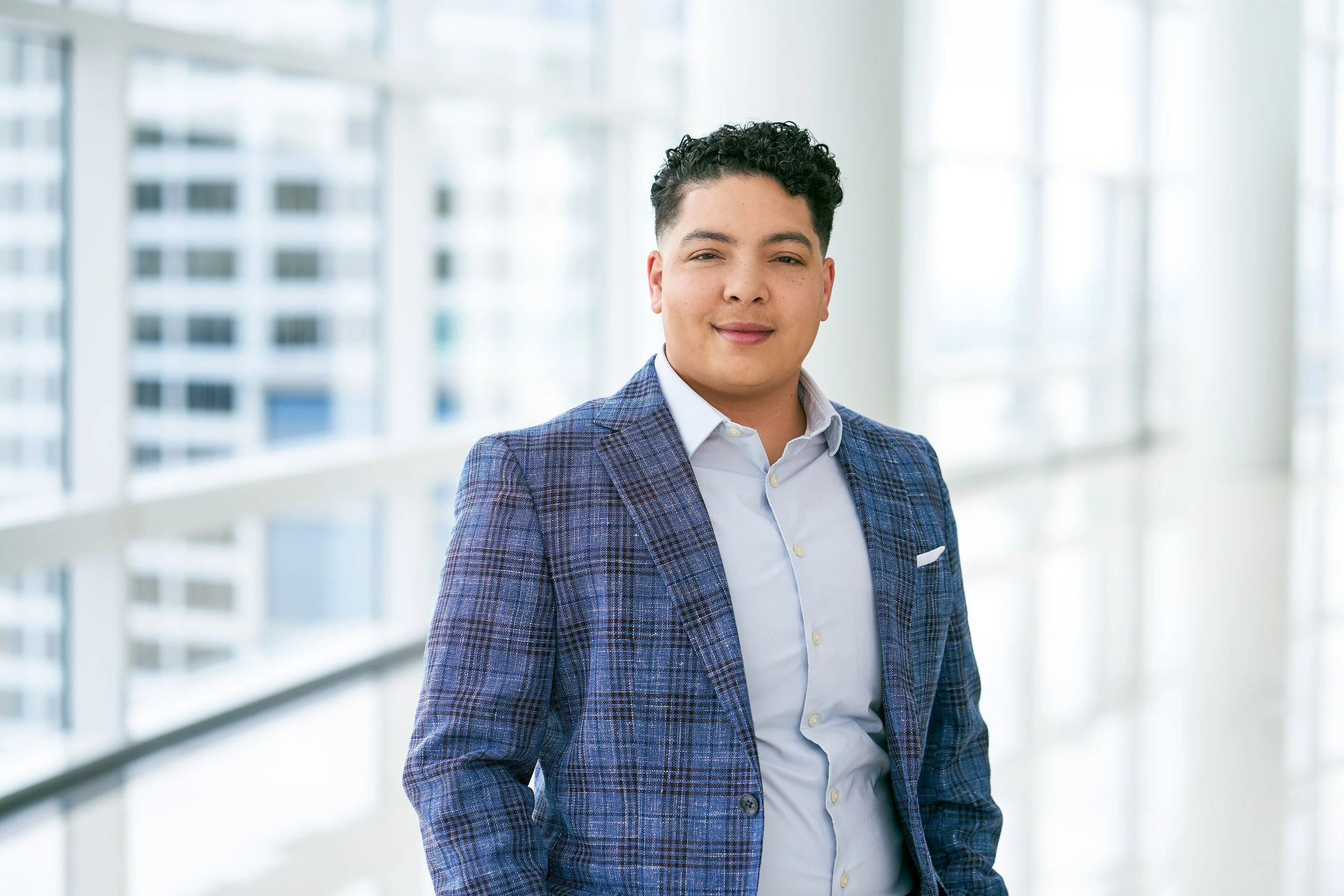 Executive portrait of business leader in blue plaid jacket standing in modern office corridor with natural window light.