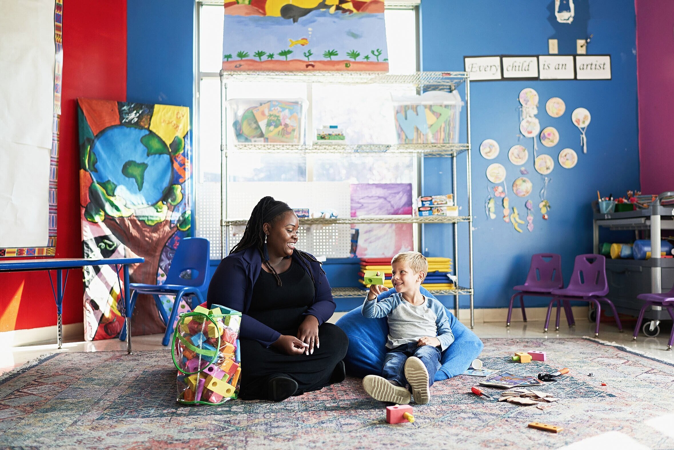 A woman and a young boy sitting on the floor of a colorful classroom, playing with building blocks and smiling at each other, surrounded by chairs, shelves, and children's artwork.