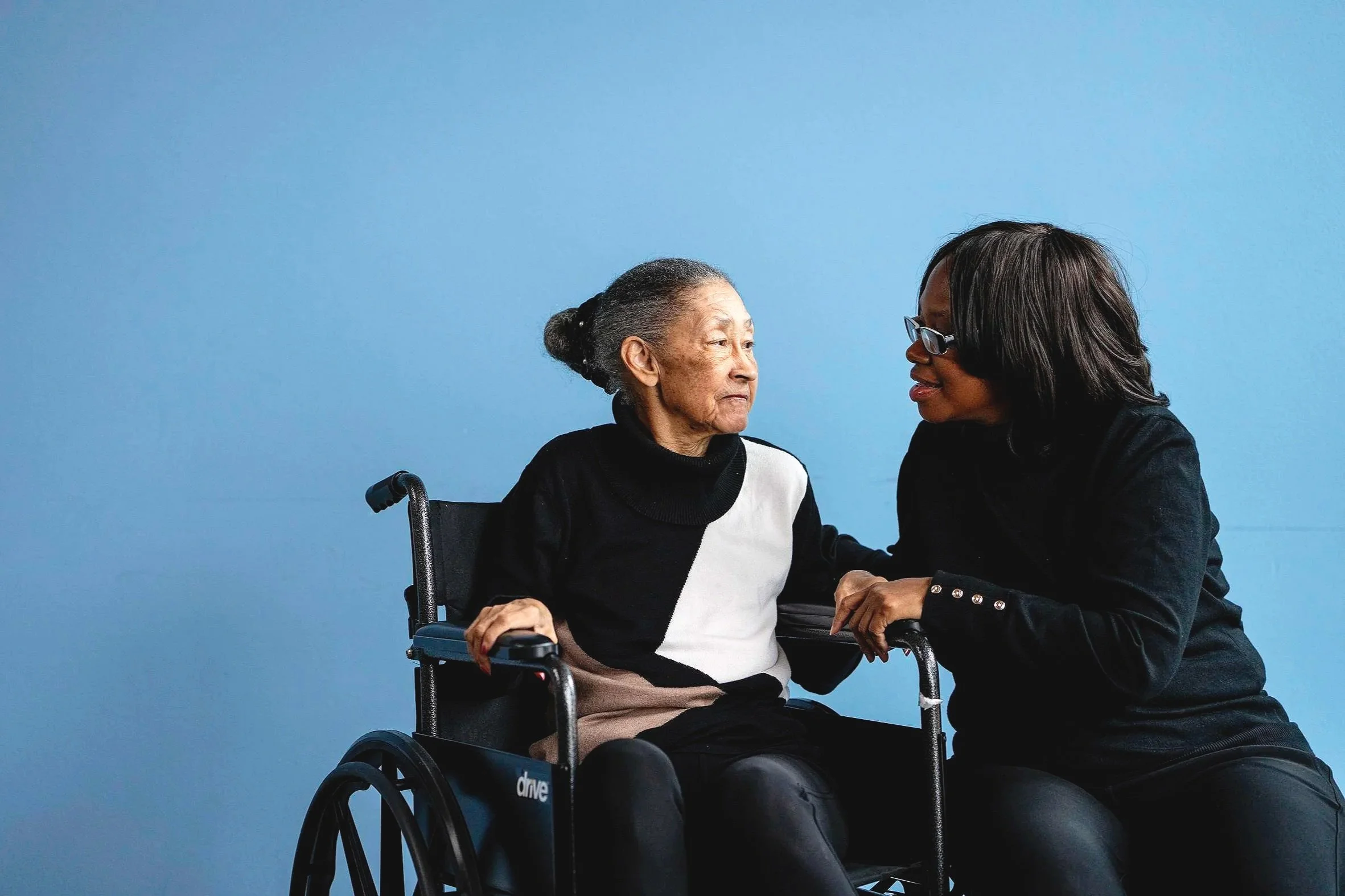 An elderly Black woman in a wheelchair shares a quiet moment with a younger Black caregiver against a soft blue background.
