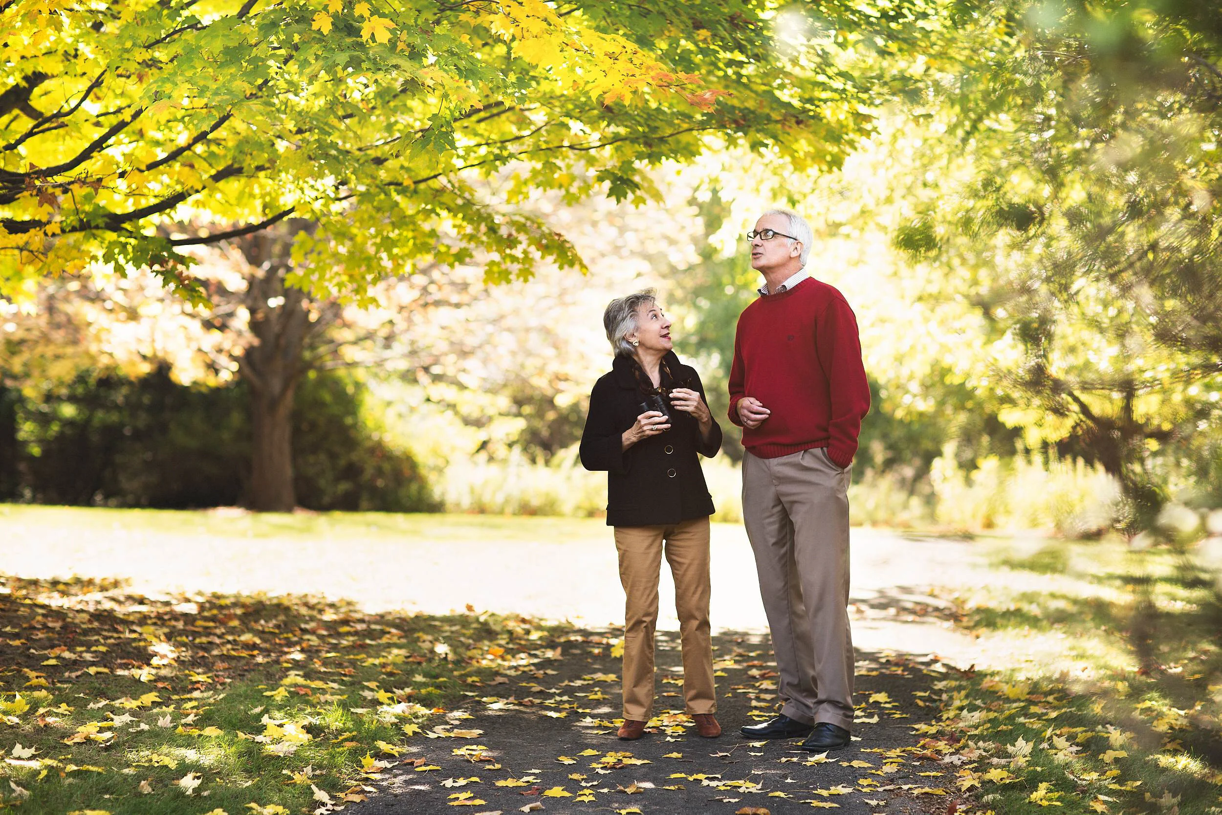An elderly man and woman talking and holding drinks while standing on a leaf-covered path in a park during fall, surrounded by trees with yellow and orange leaves.
