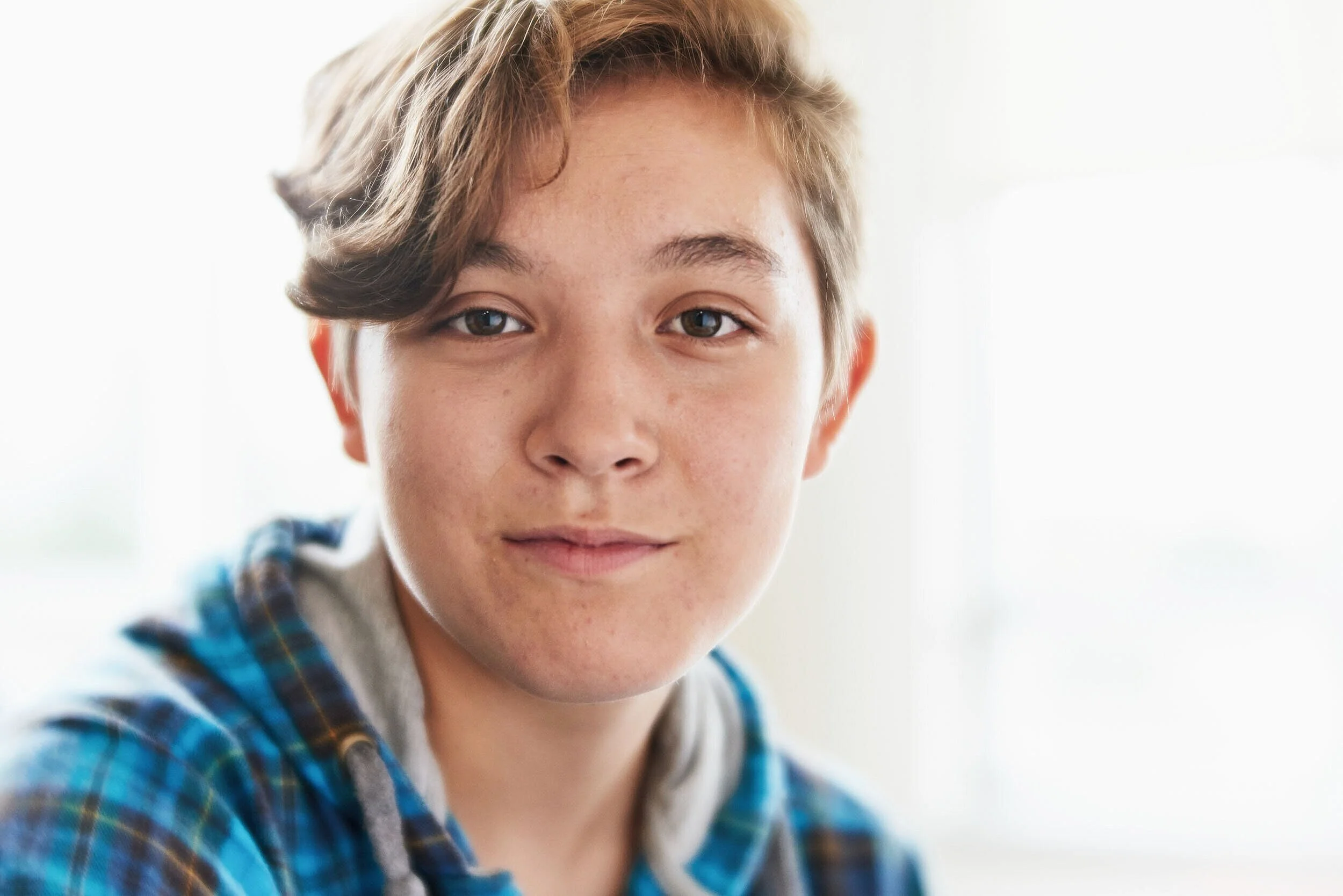 A young boy with light brown hair, tan skin, blue eyes, and a slight smile, wearing a blue and green plaid hoodie, indoors with a bright, soft-focused background.