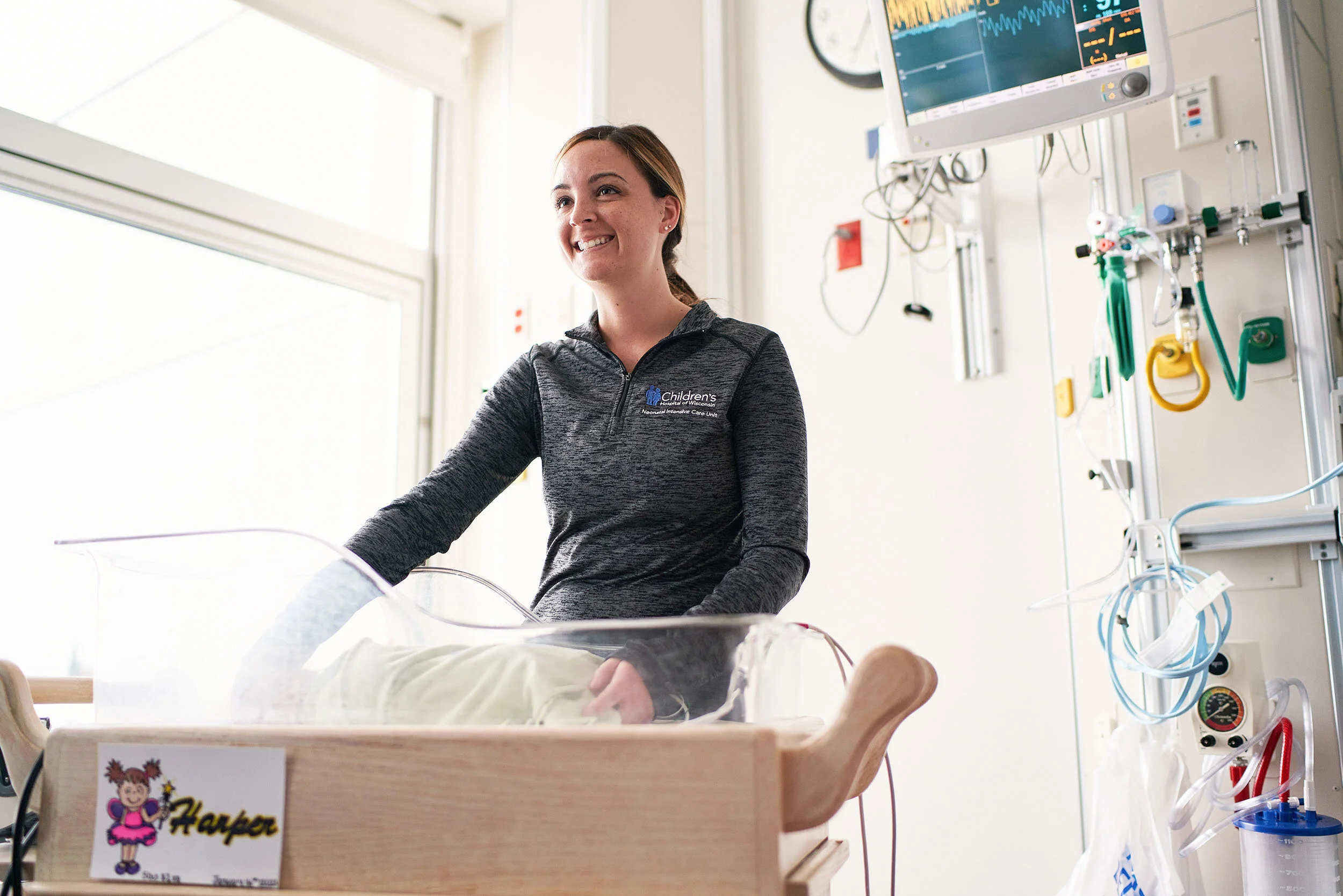 A woman smiling in a hospital NICU room standing beside a clear bassinet with a baby inside. Hospital medical equipment and monitors are visible on the wall behind her, with a logo on her sweatshirt indicating she works at a children's hospital.