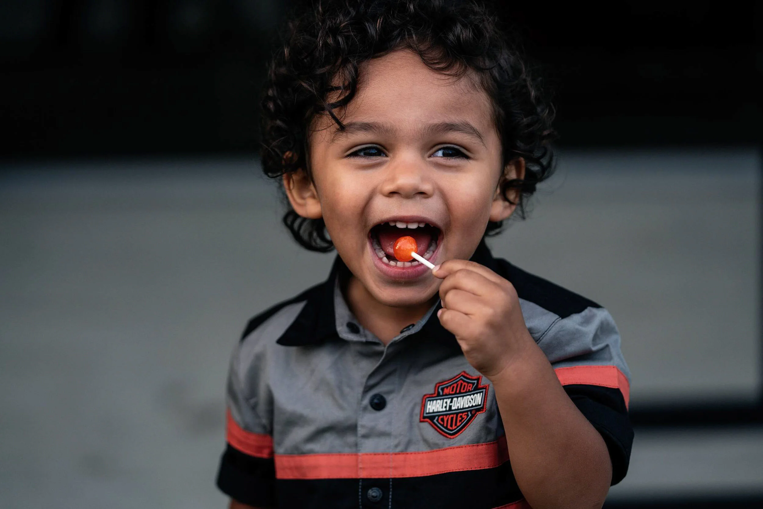 Young boy with curly hair smiling and holding a red lollipop in his mouth, wearing a gray and black Harley-Davidson shirt.