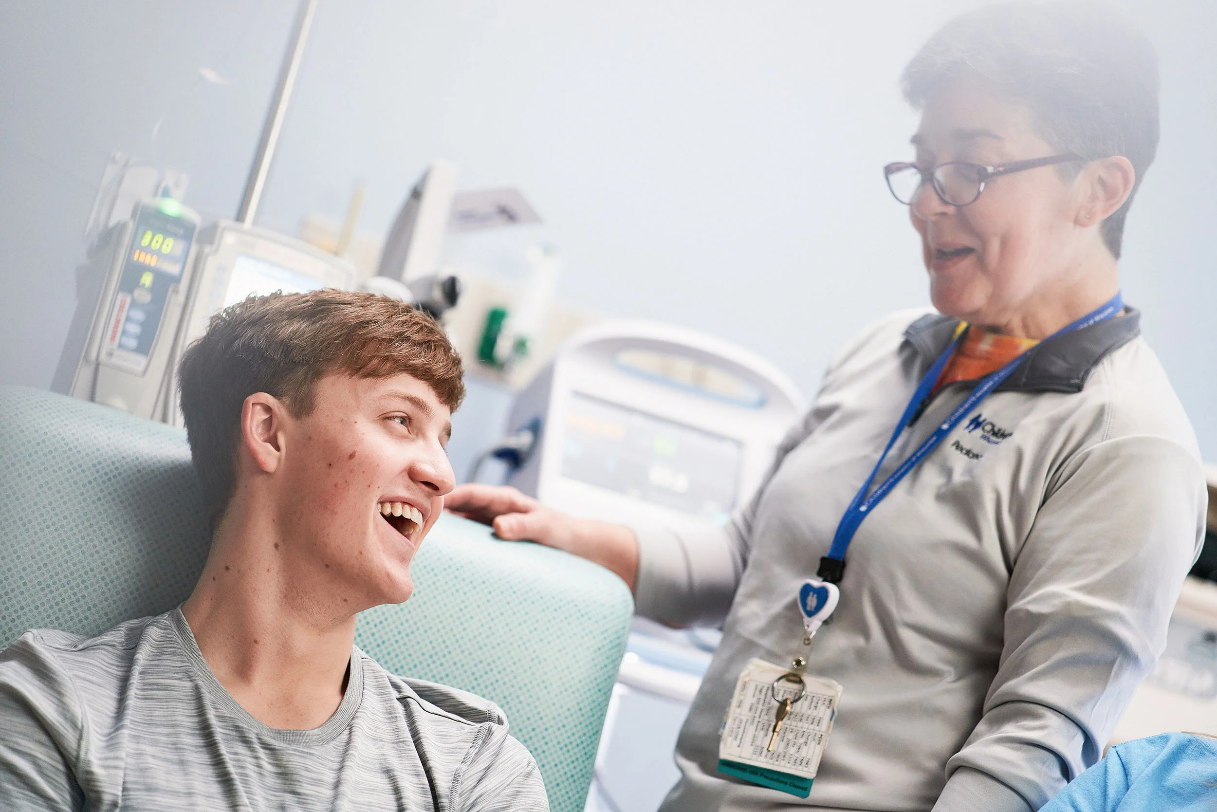 Young male patient smiling and talking with nurse in hospital room, medical equipment in background.