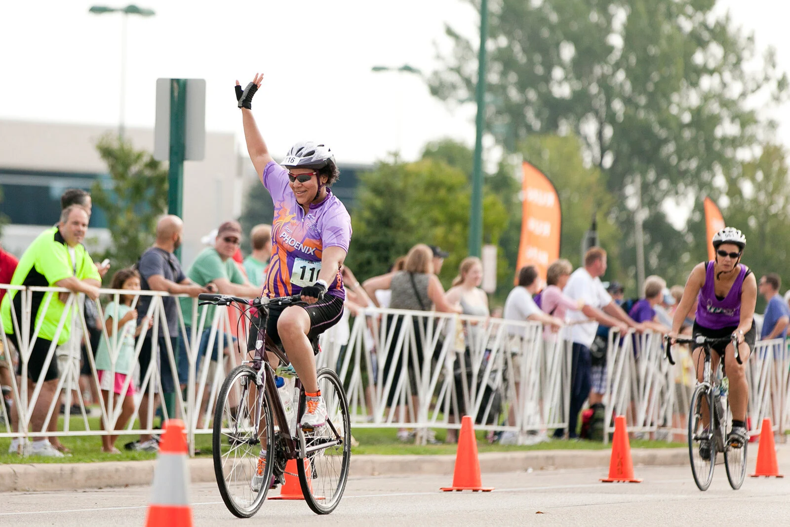 A woman in a purple cycling jersey and black shorts is riding a bike and waving at the finish line of a race, smiling, with a crowd of spectators behind her along the fence and orange cones on the road.