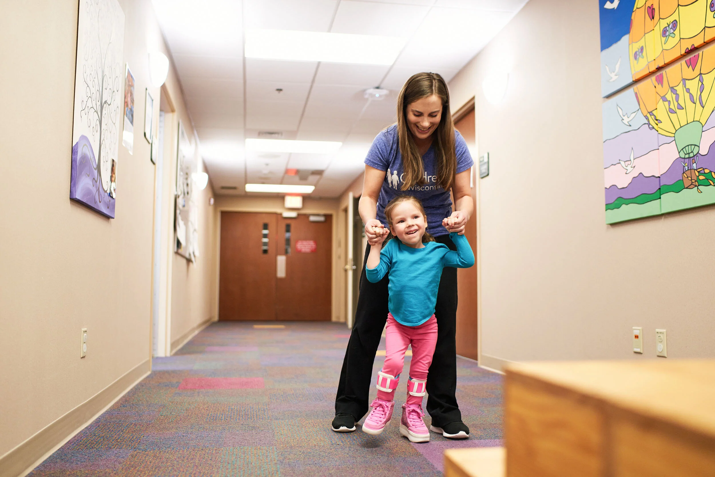 A woman is helping a young girl walk down a colorful hallway with artwork on the walls, both smiling happily.