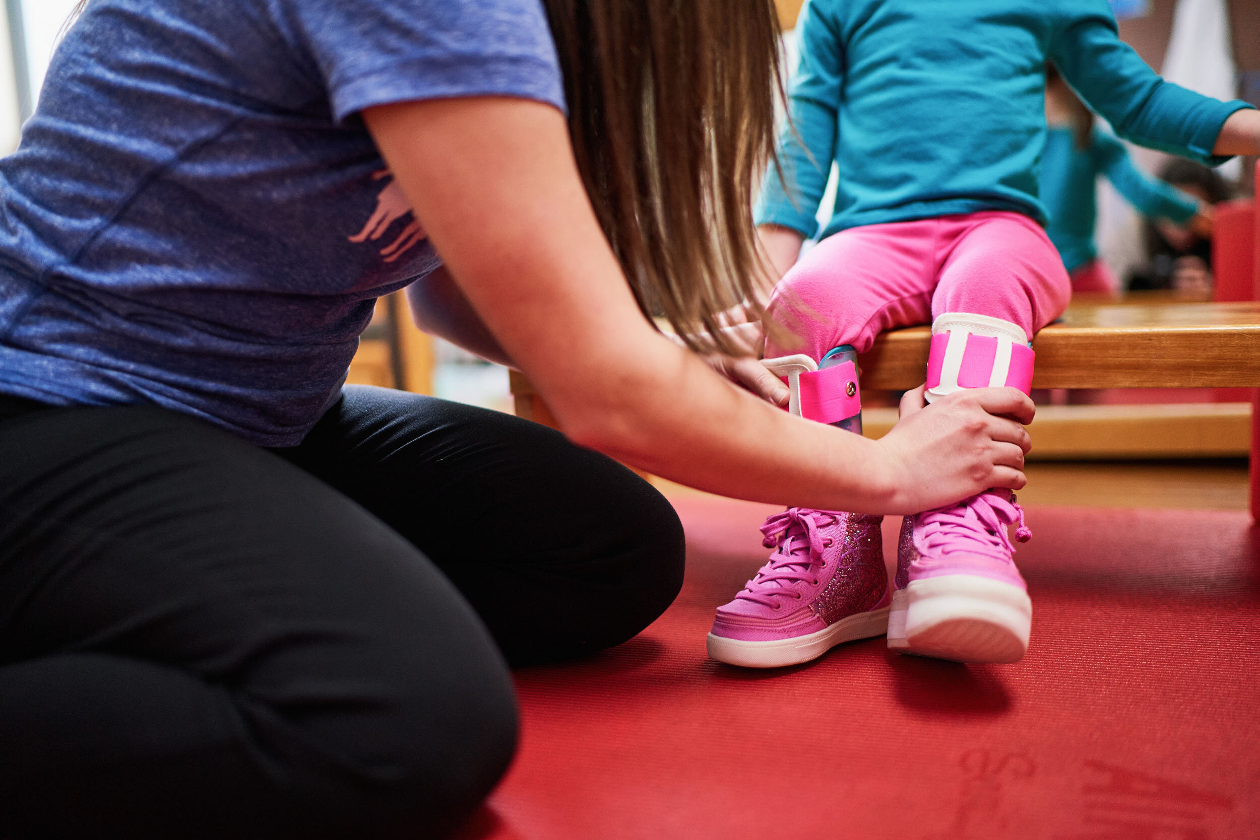 A person helping a child put on pink glittery high-top sneakers while the child sits on a wooden bench, with a red floor and other children in the background.