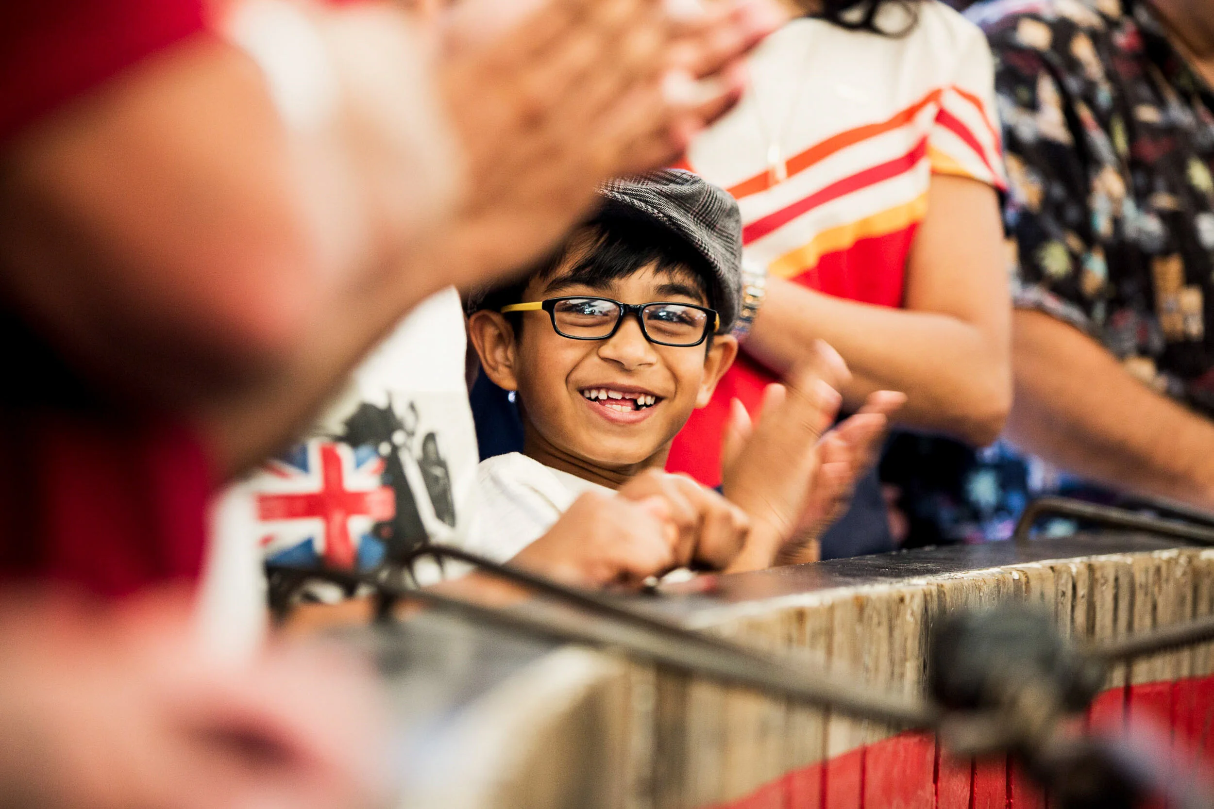 Smiling young boy with glasses and a cap claps, surrounded by adults at a festive event.