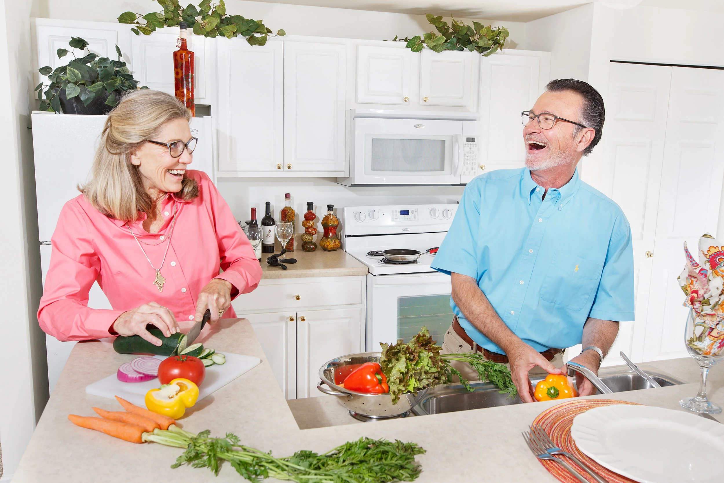 An elderly woman and man laughing and cooking together in a kitchen. The woman is slicing a cucumber while the man holds a yellow bell pepper. There are vegetables, including carrots, tomatoes, and lettuce, on the counter. The kitchen has white cabin