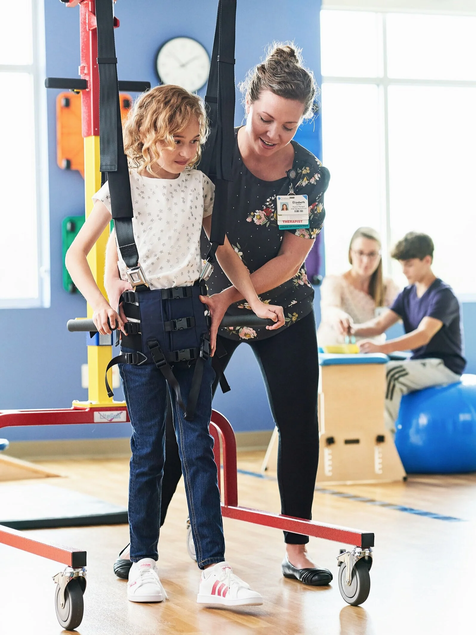 A therapy session at a rehabilitation center where a woman therapist is assisting a young girl with a harness and support equipment, with other children in the background engaging in activities.