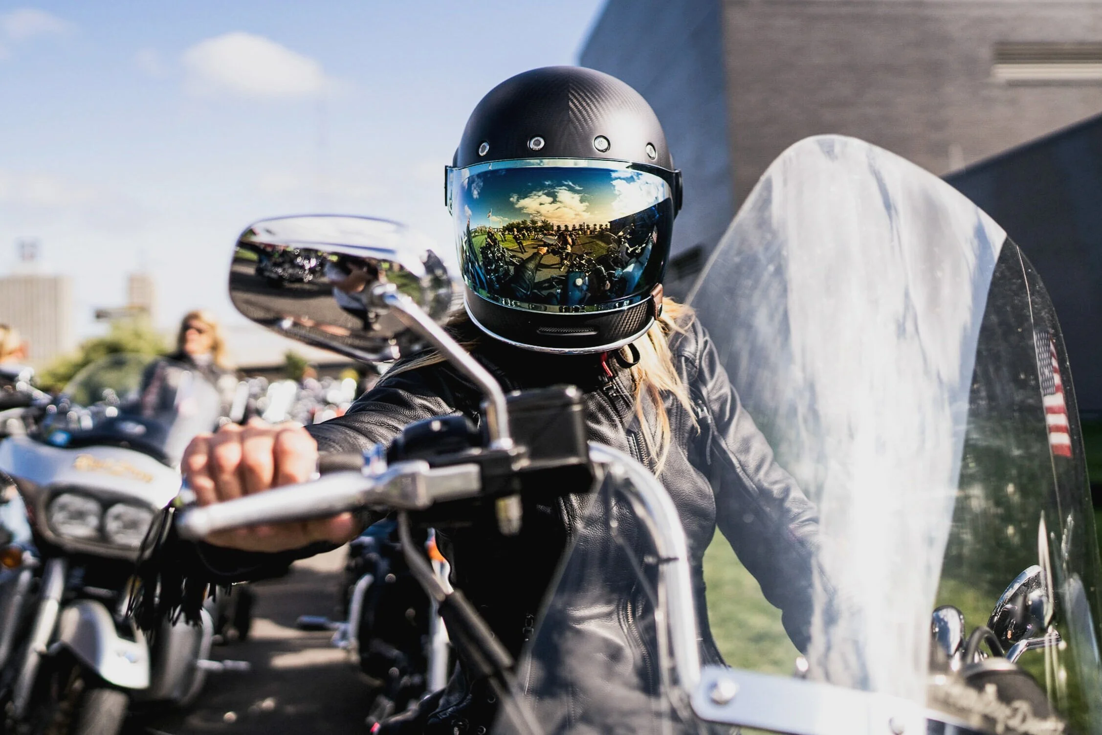 Person wearing a black helmet and leather jacket riding a motorcycle with a clear windshield, surrounded by others at a motorcycle gathering on a sunny day.