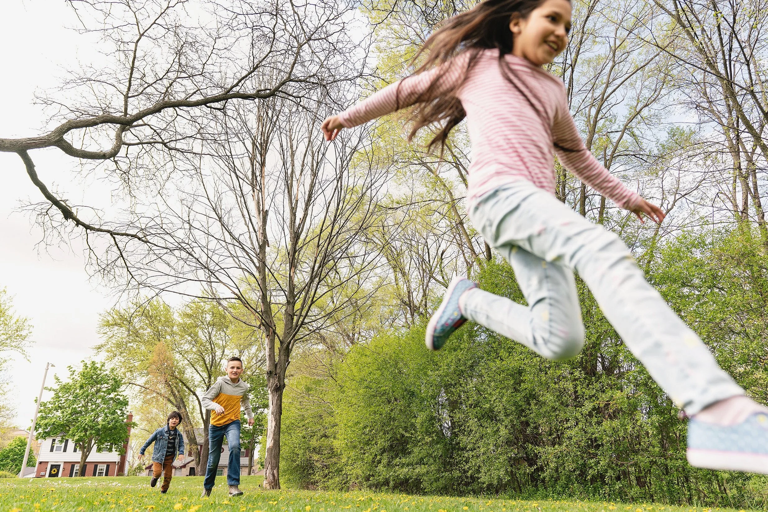 Three children playing and running in a grassy park with trees