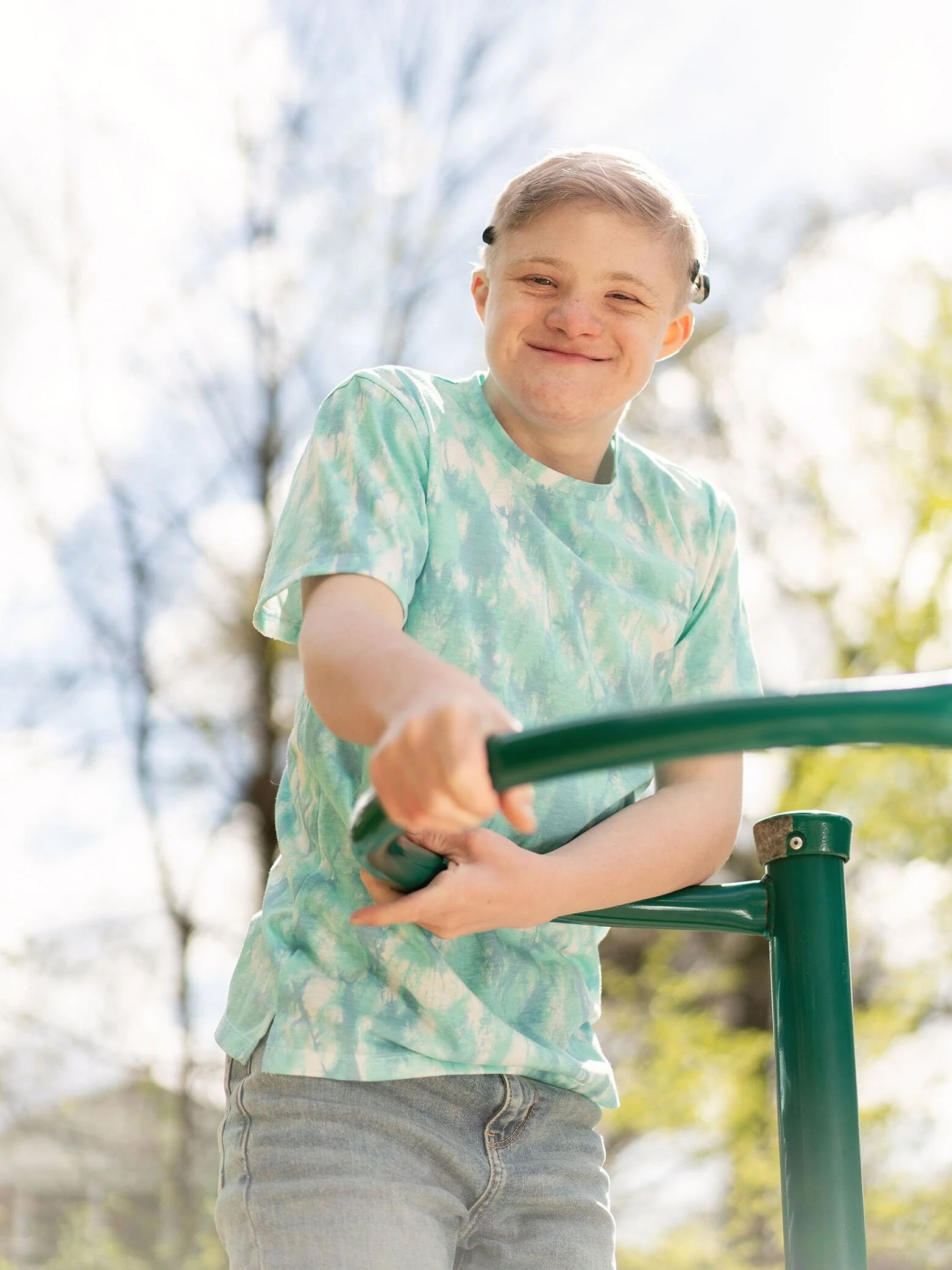 Young boy smiling while holding onto a green playground bar outdoors on a sunny day.