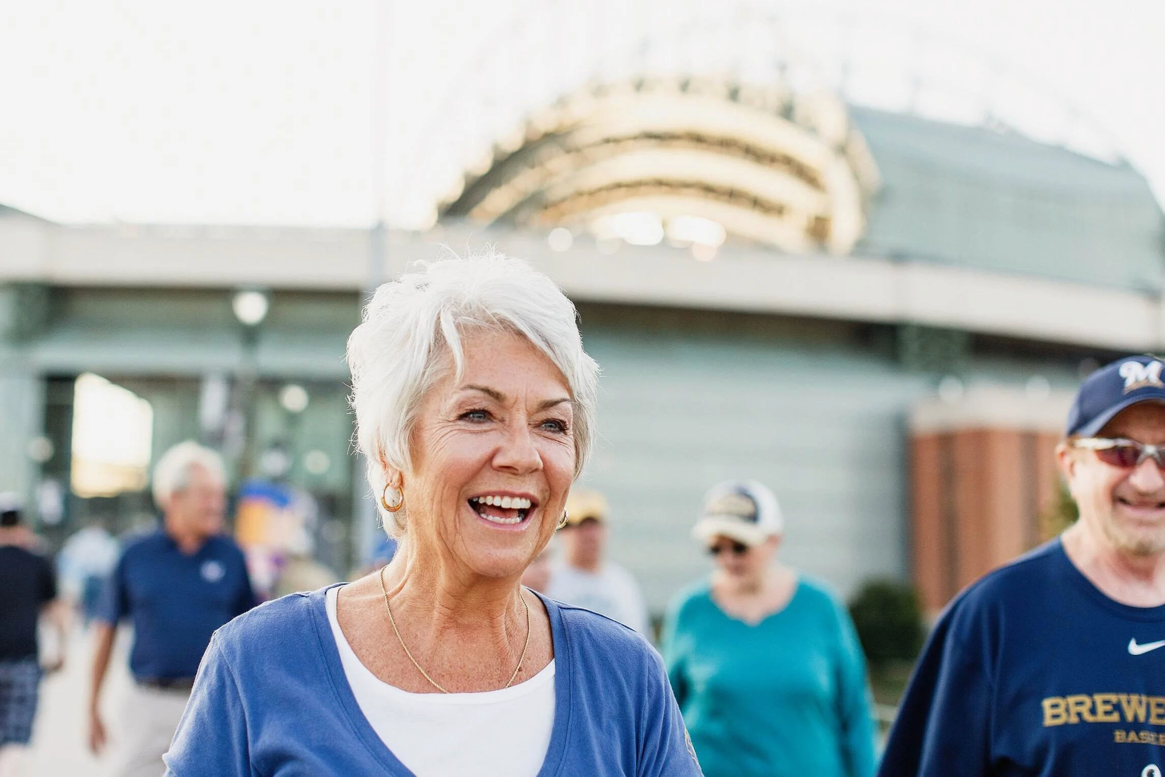 An elderly woman with short white hair and a blue shirt, smiling outdoors among other people, with a large building in the background.